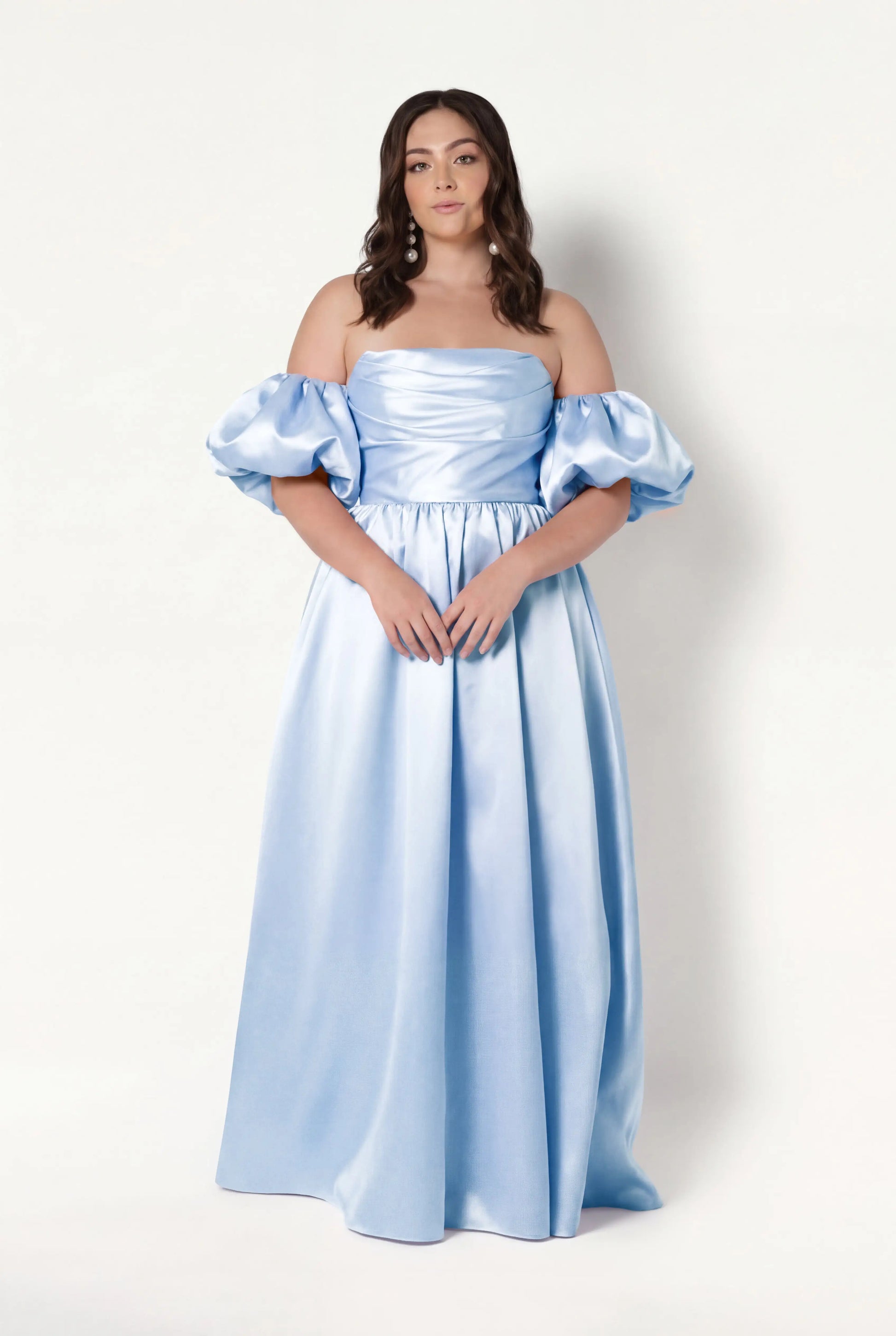 A woman with wavy brown hair and a neutral expression models the TH&TH Blair Bridesmaid Dress in Frosted Blue Satin Twill with detachable puff sleeves, standing against a plain white background.