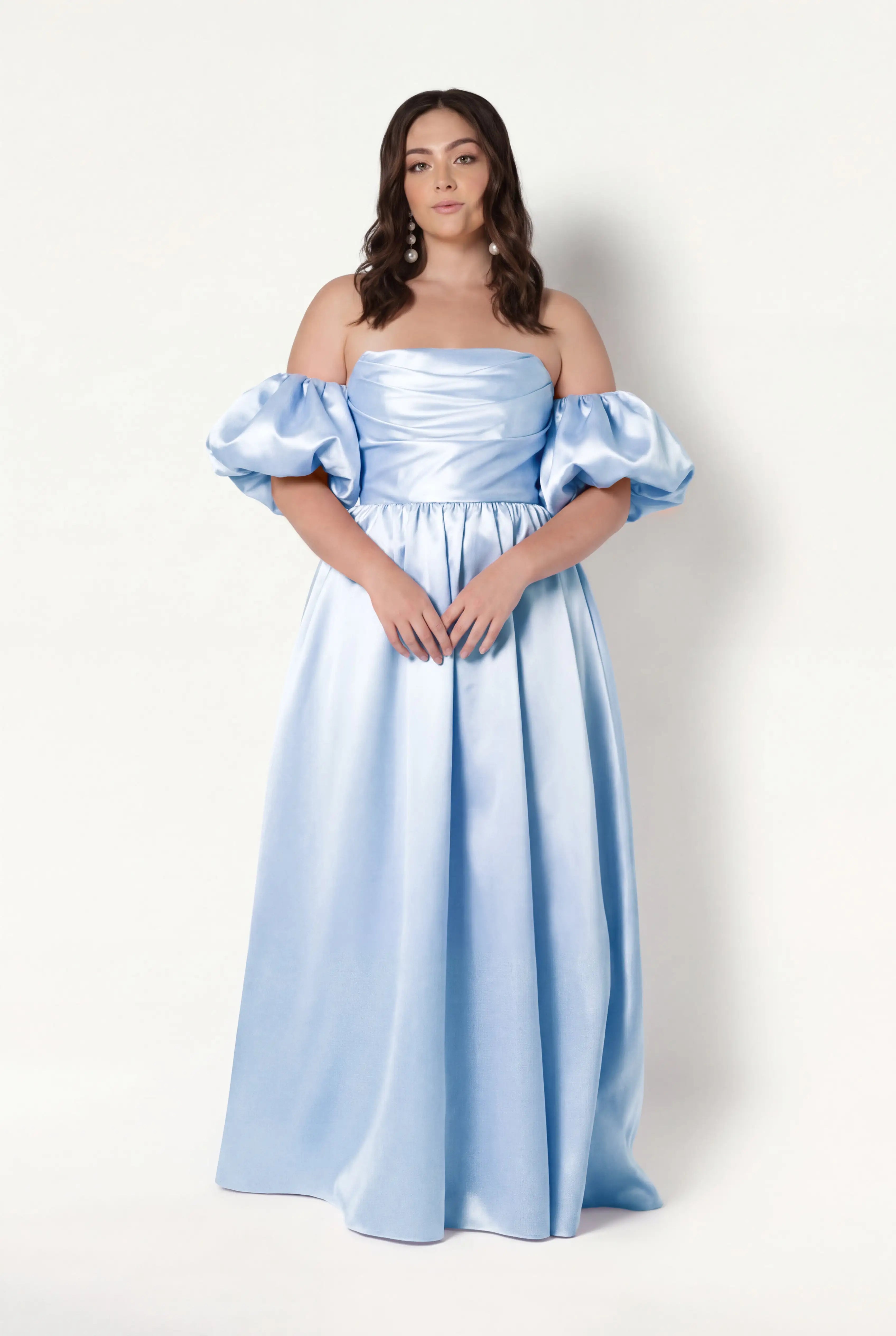 A woman with wavy brown hair and a neutral expression models the TH&TH Blair Bridesmaid Dress in Frosted Blue Satin Twill with detachable puff sleeves, standing against a plain white background.