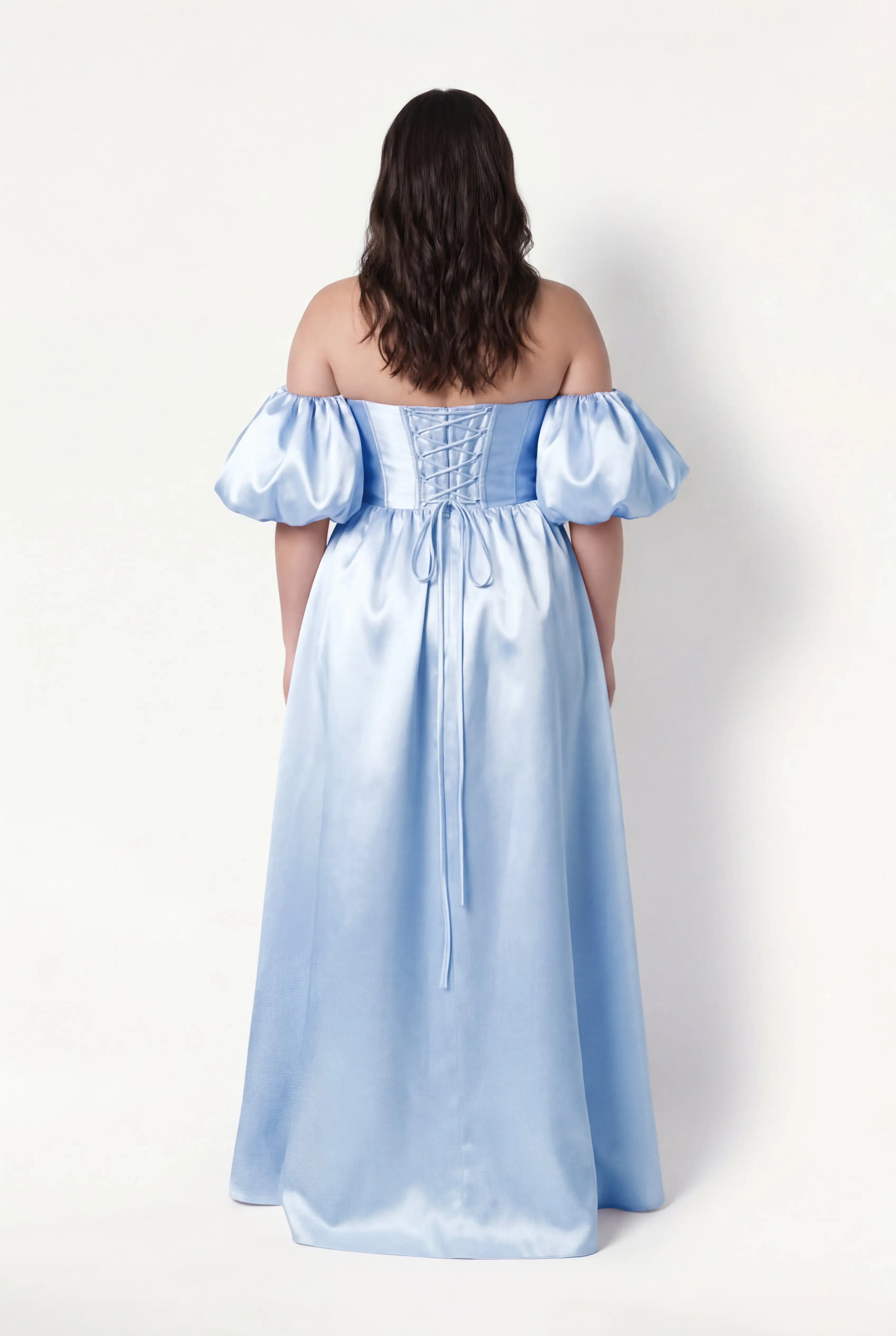 A woman with wavy brown hair stands with her back turned, wearing the TH&TH Blair Bridesmaid Dress in Frosted Blue Satin Twill, featuring an off-the-shoulder design and detachable puff sleeves, against a plain white background.
