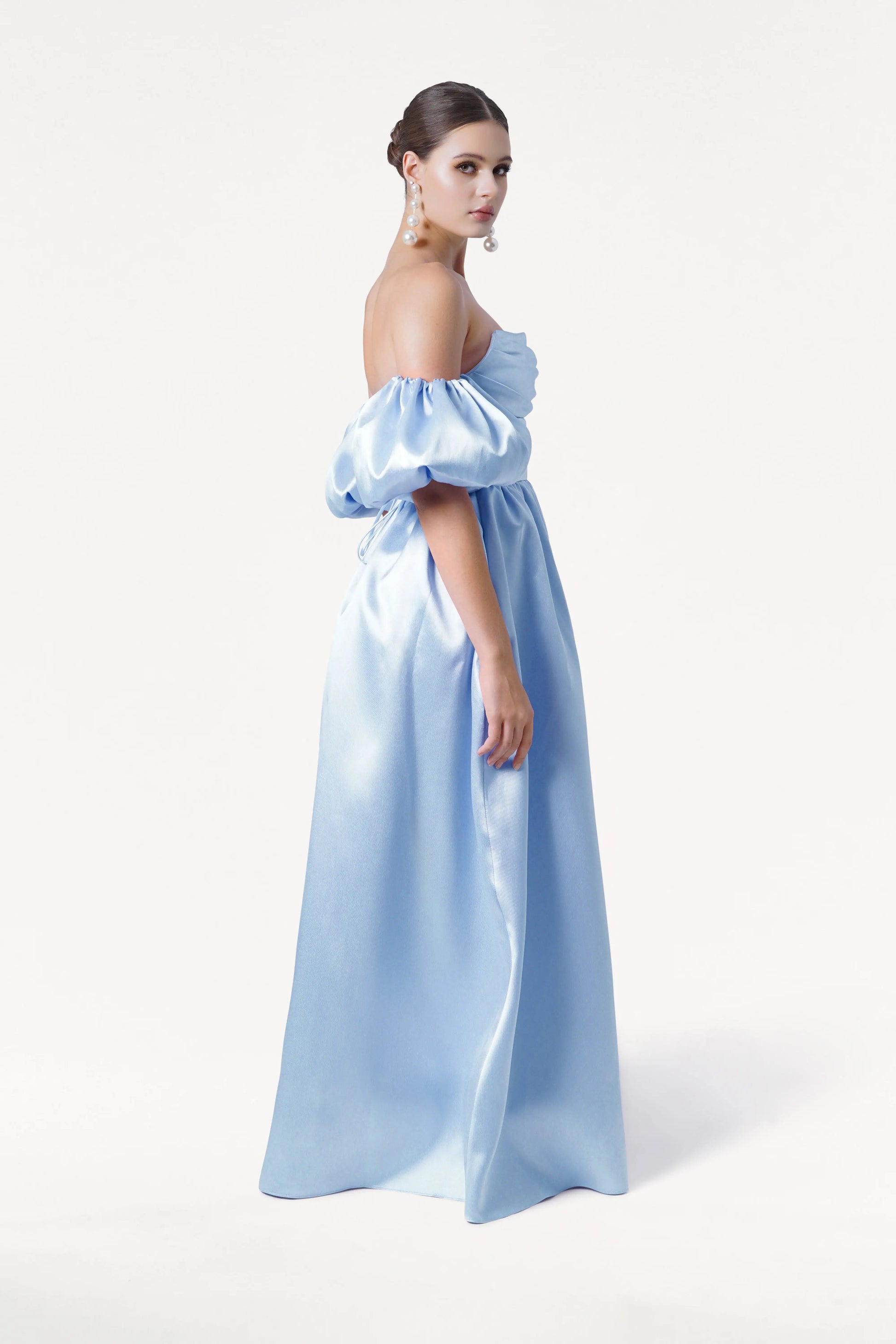 A woman stands sideways in the Blair Bridesmaid Dress in Frosted Blue Satin Twill by TH&TH, featuring an off-the-shoulder corset bodice and detachable puff sleeves. She wears pearl earrings with her hair in a sleek bun against a white background.