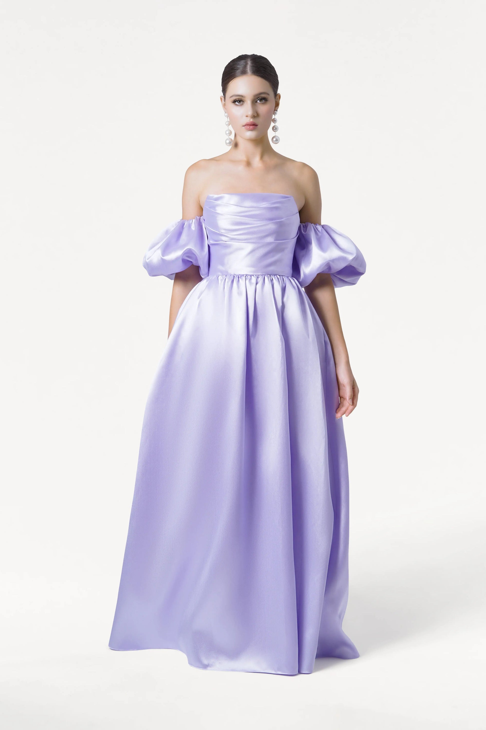 A woman wears the TH&TH Blair Bridesmaid Dress in Lilac Mist Satin Twill, featuring an off-the-shoulder neckline, voluminous puff sleeves, a full skirt, and corset bodice. Her hair is pulled back with statement earrings against a plain white background.