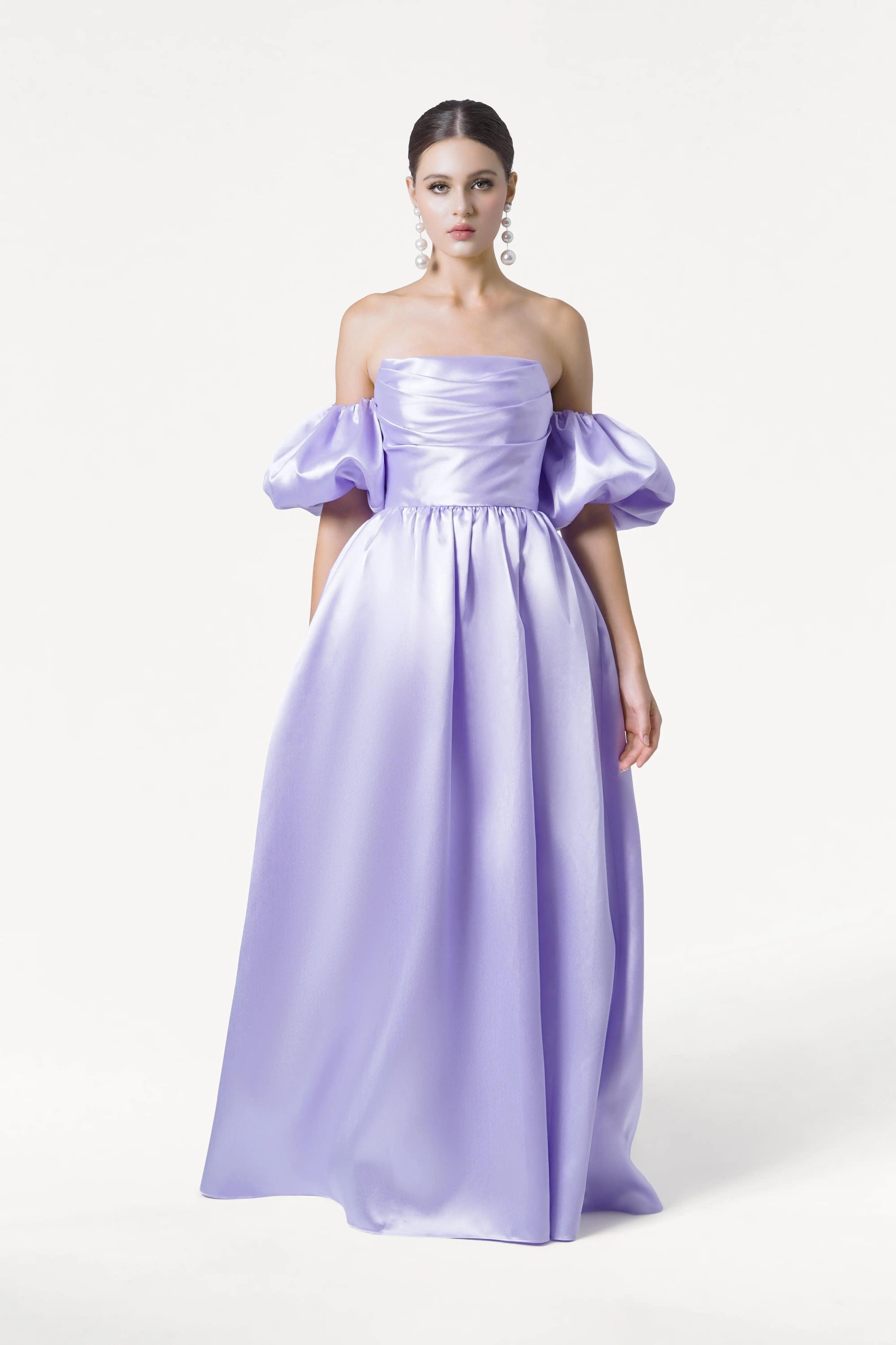 A woman wears the TH&TH Blair Bridesmaid Dress in Lilac Mist Satin Twill, featuring an off-the-shoulder neckline, voluminous puff sleeves, a full skirt, and corset bodice. Her hair is pulled back with statement earrings against a plain white background.