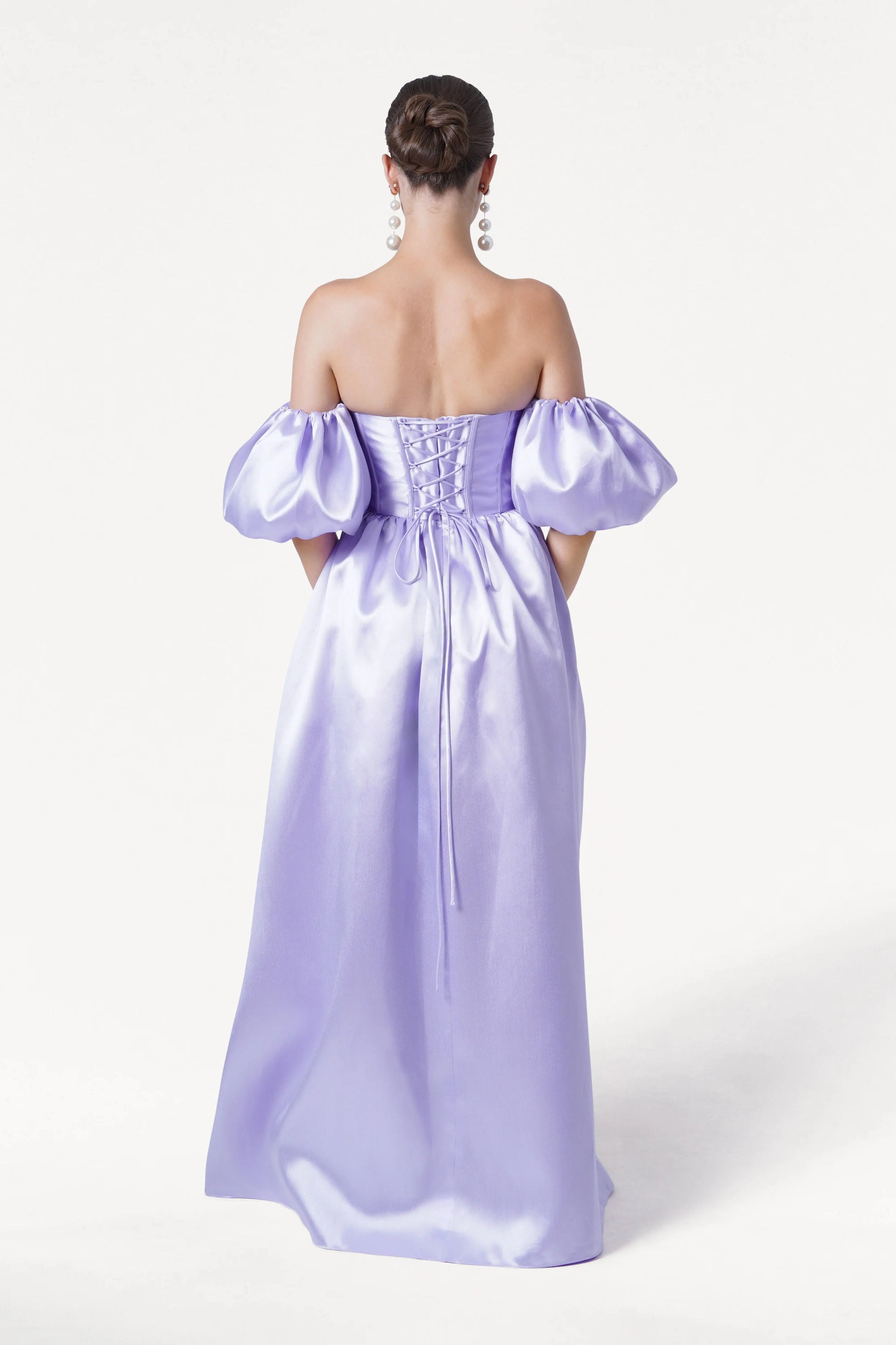 A woman stands with her back to the camera, wearing the TH&TH Blair Bridesmaid Dress in Lilac Mist Satin Twill—a gown with a corset bodice, puffed sleeves, and full-length skirt. Her hair is styled in a sleek updo.