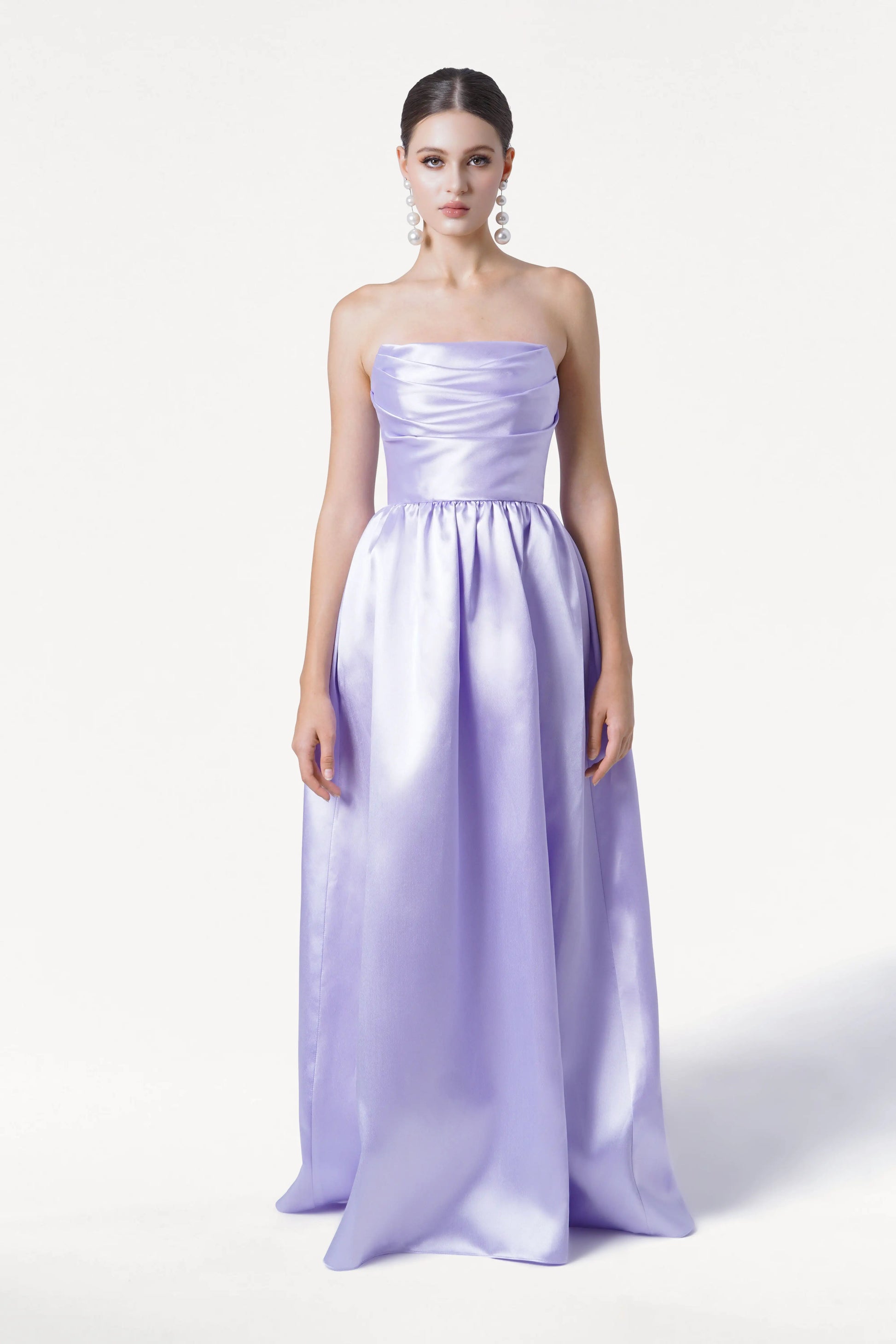 A woman stands against a white background in the TH&TH Blair Bridesmaid Dress in Lilac Mist Satin Twill—a strapless, floor-length gown with a corset bodice and full skirt. She wears pearl drop earrings and has her dark hair styled back.