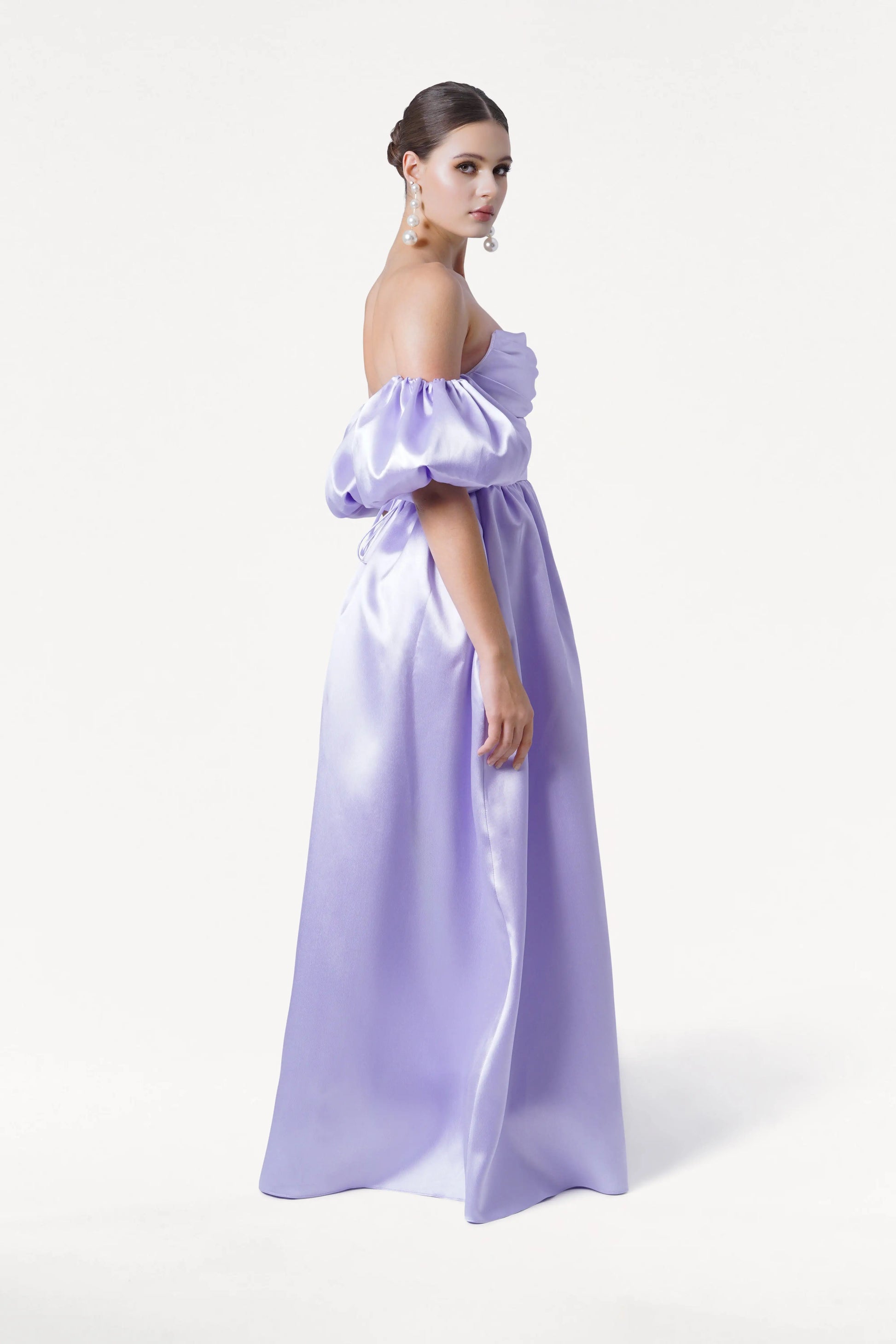 A woman stands sideways against a white background, wearing the TH&TH Blair Bridesmaid Dress in Lilac Mist Satin Twill. Her hair is styled in an elegant updo with statement earrings, highlighting the dress's off-the-shoulder puffy sleeves and corset bodice.