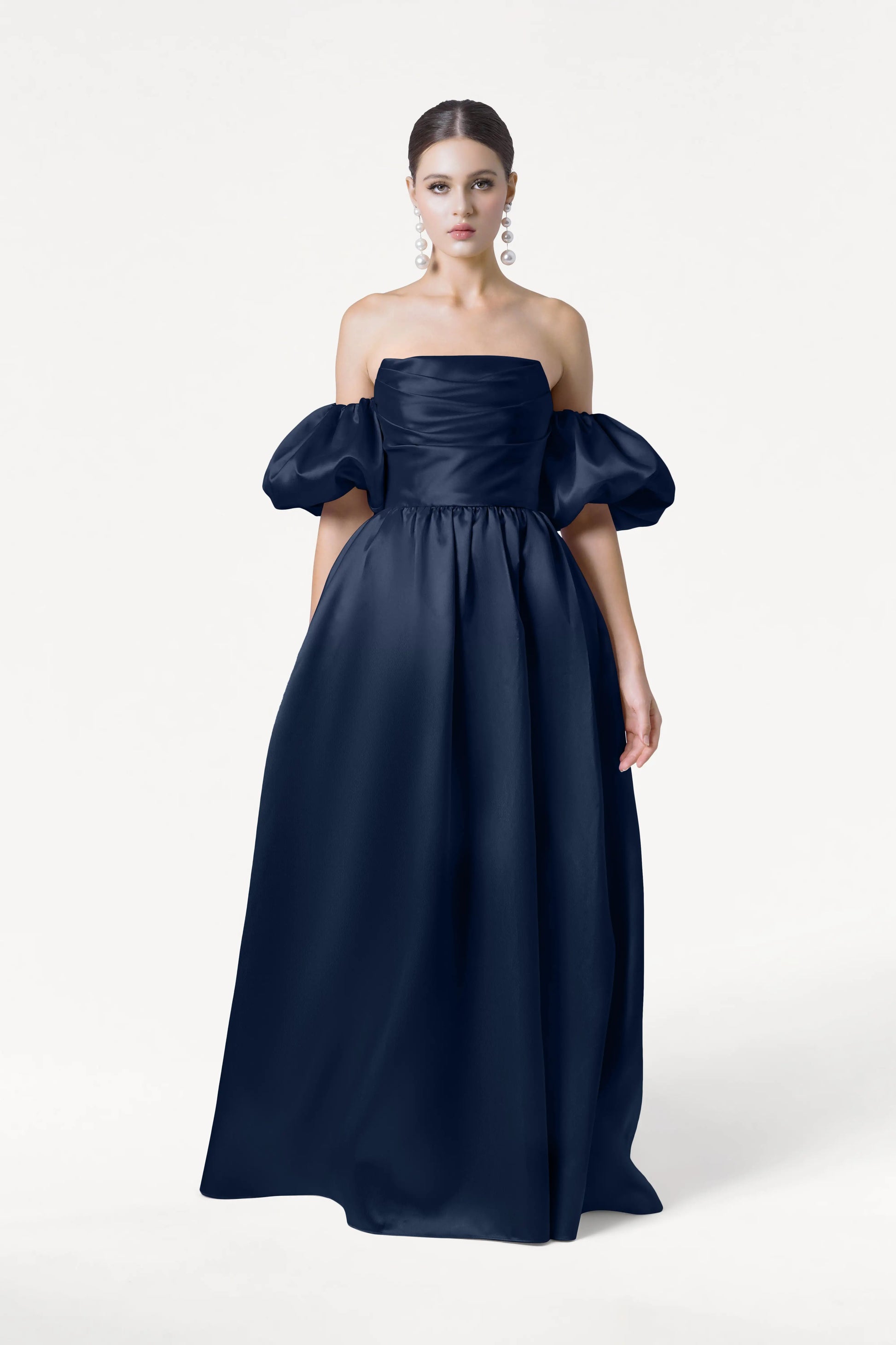 A woman in the Blair Bridesmaid Dress by TH&TH, featuring navy satin twill, off-the-shoulder design, detachable puff sleeves, and a full skirt, poses with large dangling earrings against a plain white background.