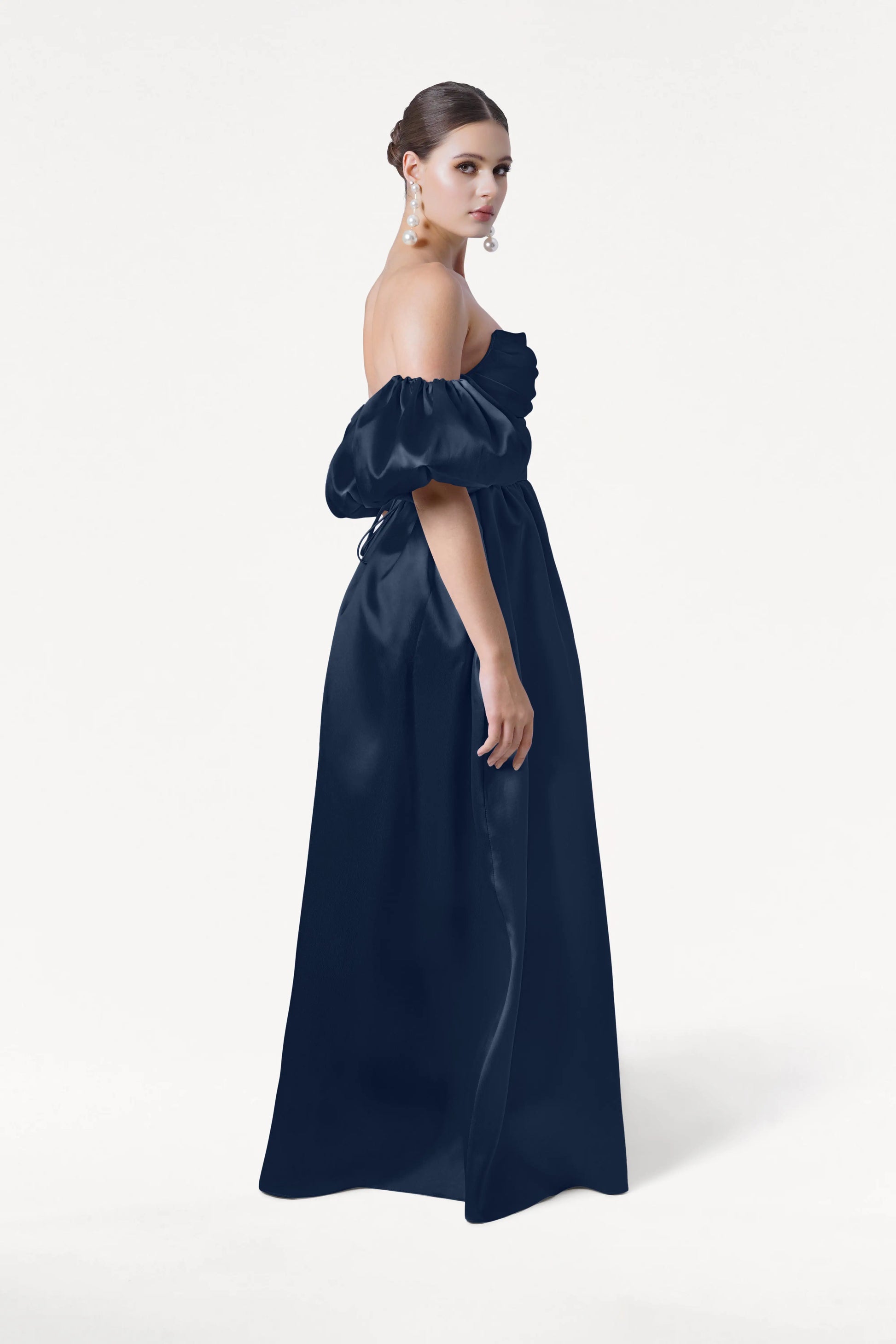 A woman in profile wears the Blair Bridesmaid Dress in Navy Satin Twill by TH&TH, featuring detachable puff sleeves. Her hair is in a low bun with large pearl earrings, set against a plain white background.
