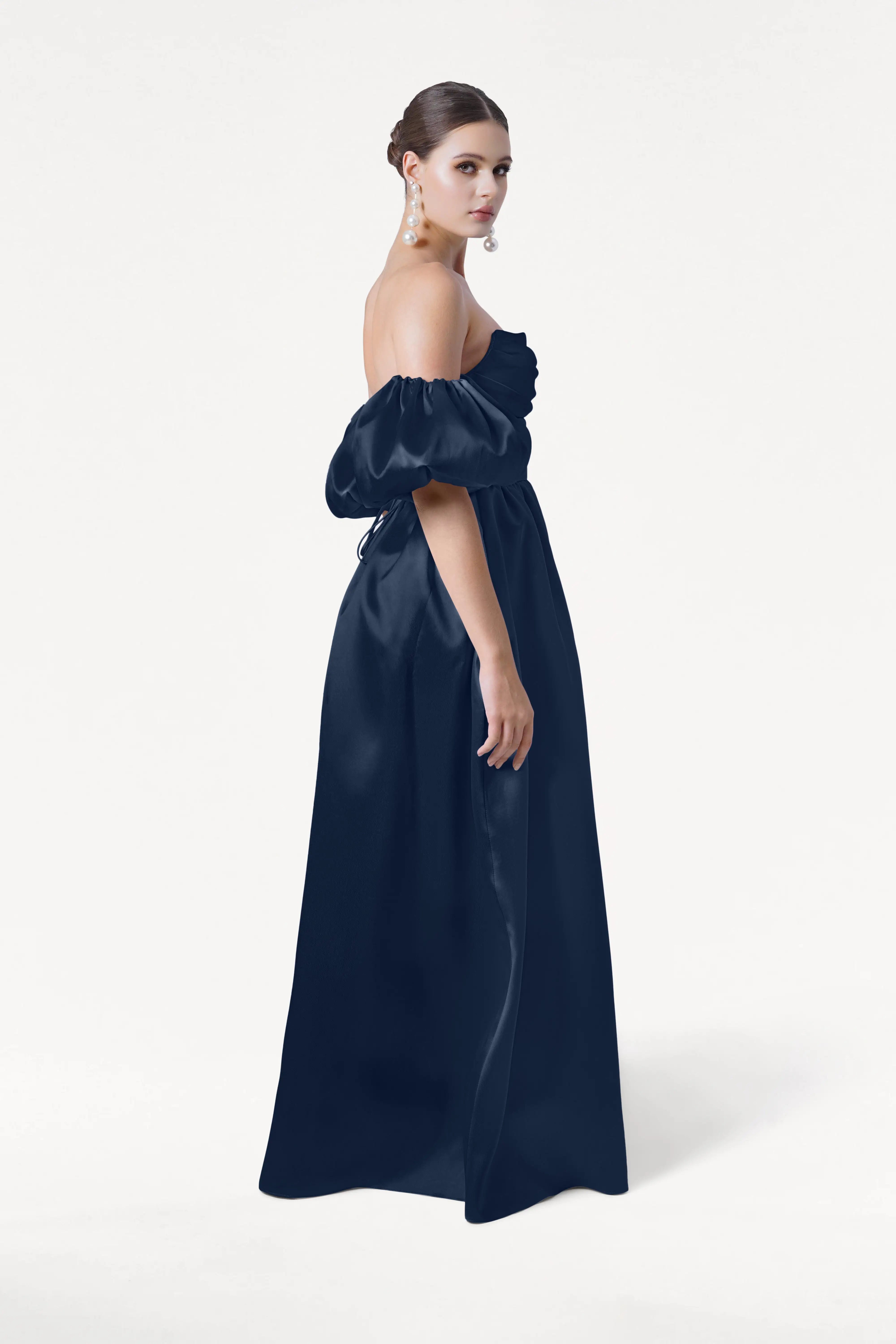 A woman in profile wears the Blair Bridesmaid Dress in Navy Satin Twill by TH&TH, featuring detachable puff sleeves. Her hair is in a low bun with large pearl earrings, set against a plain white background.