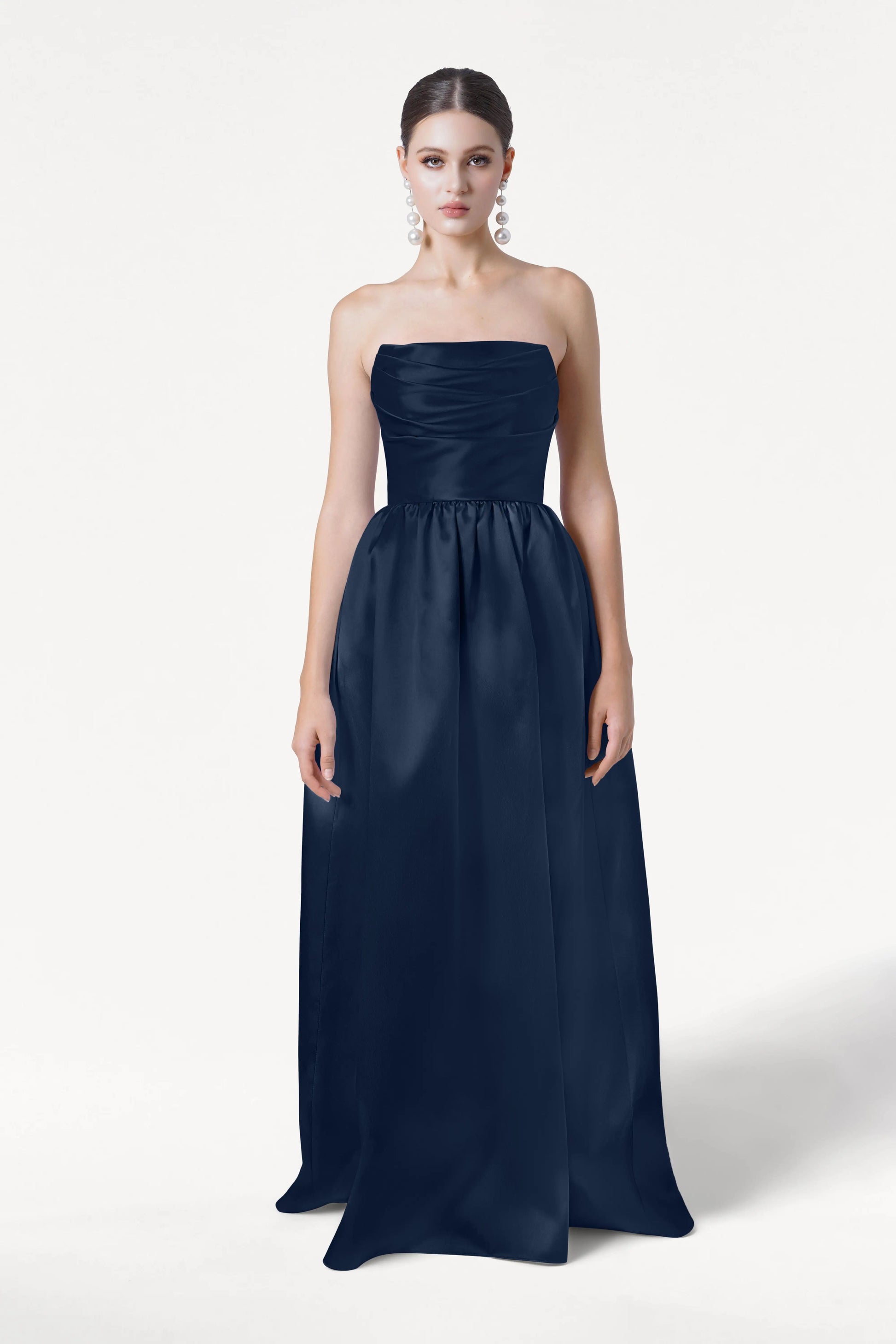 A woman stands against a plain white background wearing the Blair Bridesmaid Dress in Navy Satin Twill by TH&TH, a strapless, floor-length gown with a fitted bodice and full skirt. Her hair is pulled back and she wears large, dangling earrings.