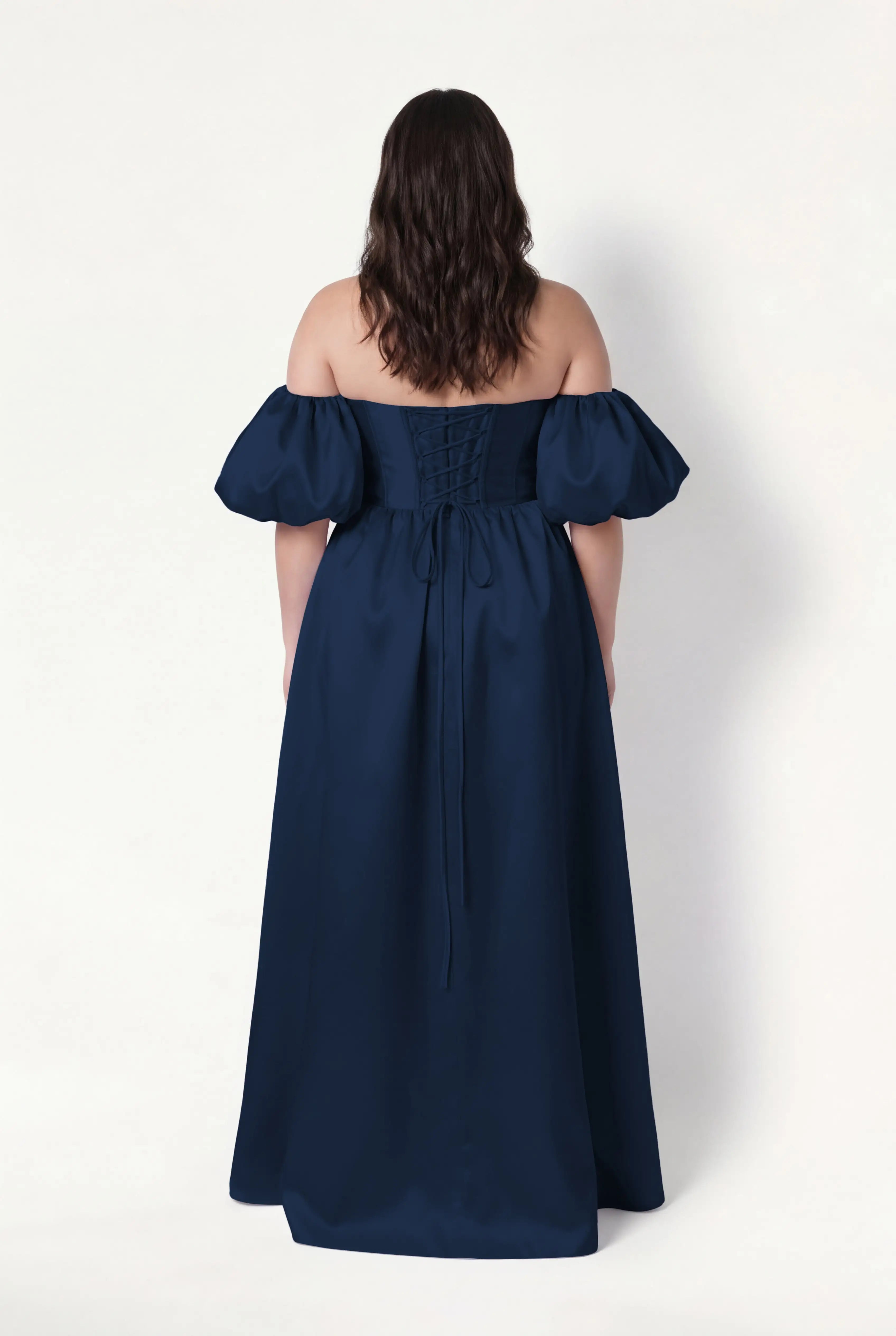 A woman with wavy brown hair wears the TH&TH Blair Bridesmaid Dress in Navy Satin Twill, featuring a floor-length silhouette, corset bodice, detachable puff sleeves, and lace-up back, standing against a plain white background facing away.