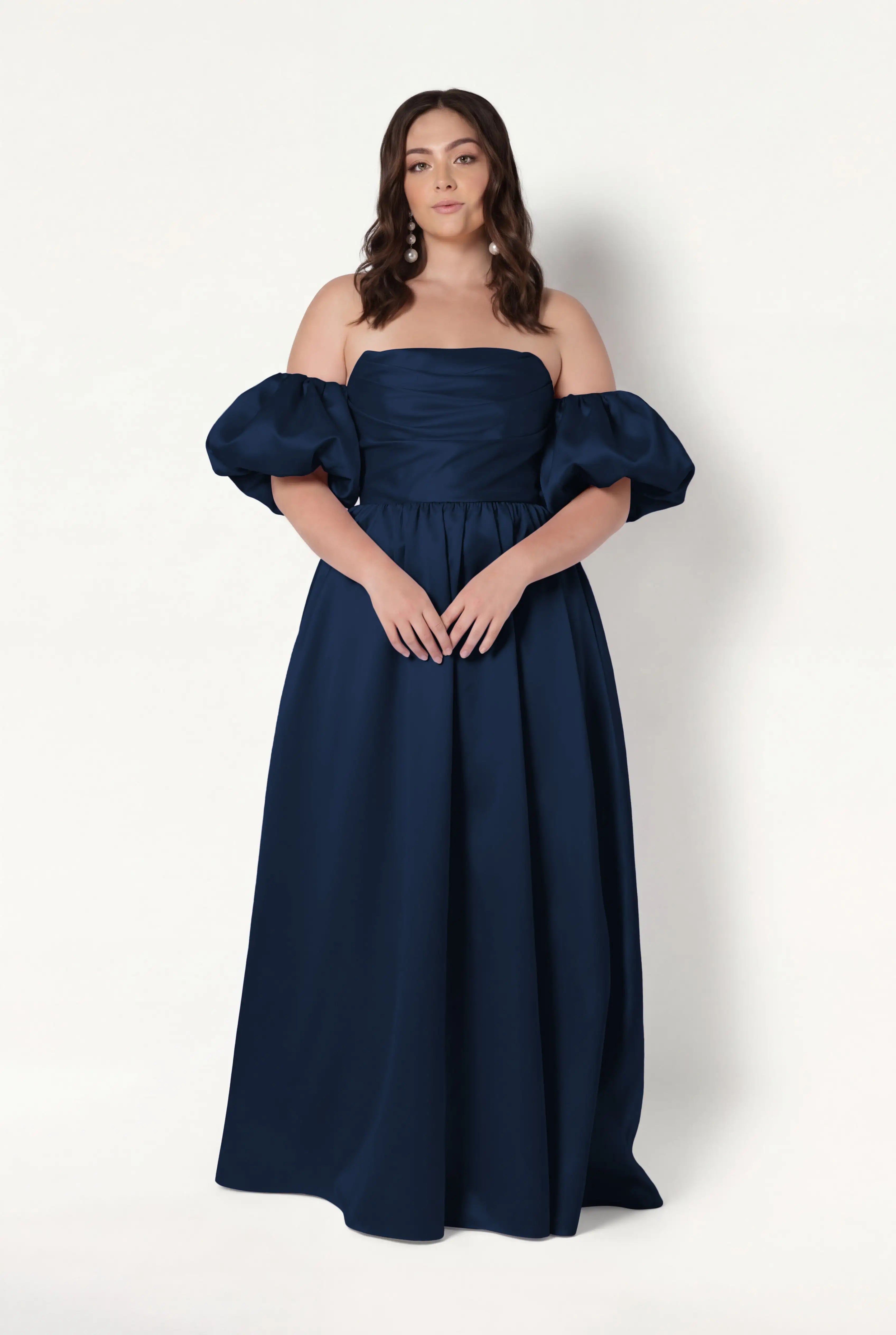A woman wears the Blair Bridesmaid Dress in Navy Satin Twill by TH&TH, featuring an off-the-shoulder corset design with detachable puff sleeves, as she stands against a plain white background, looking at the camera neutrally.
