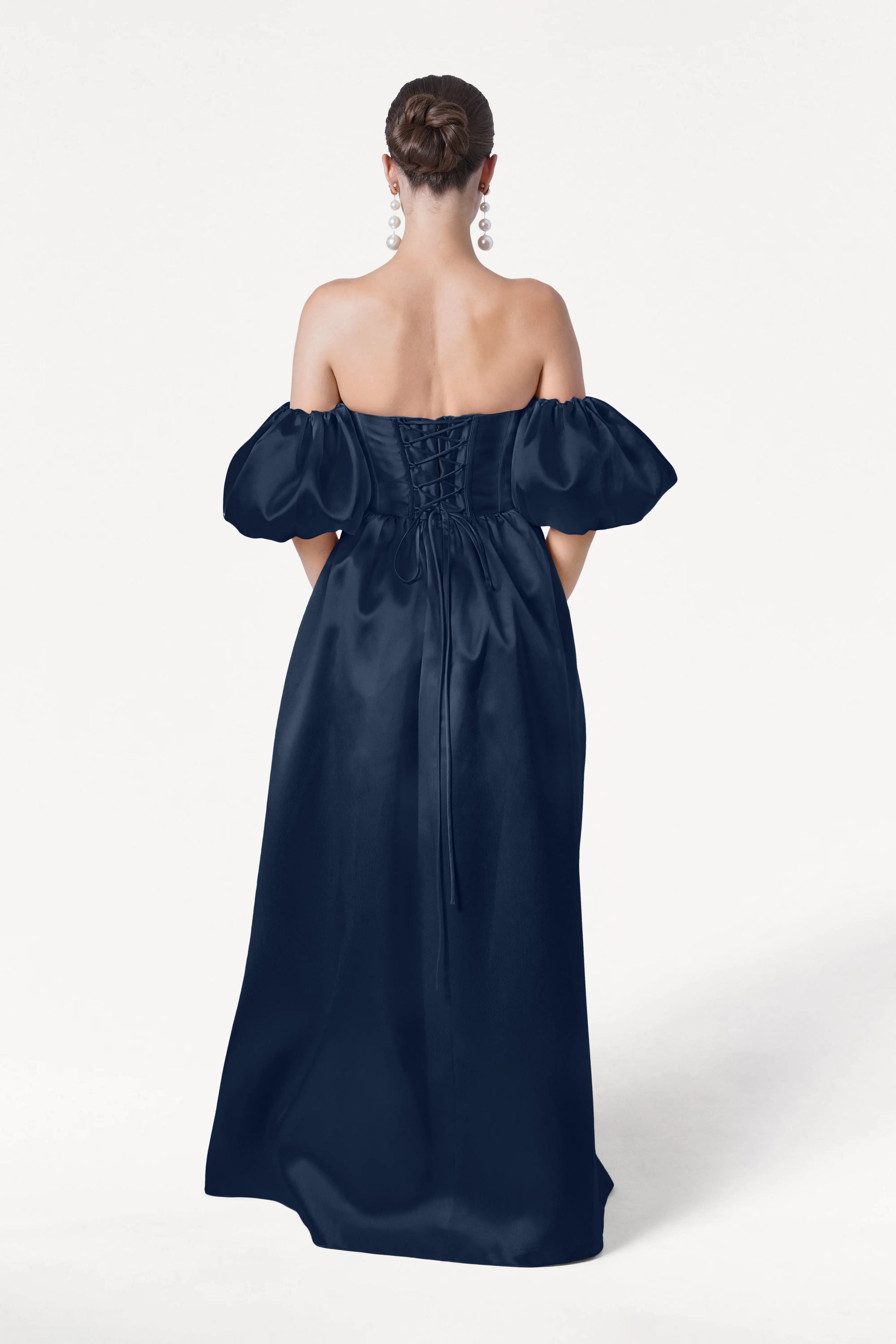 A woman with her back to the camera wears the Blair Bridesmaid Dress in Navy Satin Twill by TH&TH, featuring detachable puff sleeves and a lace-up back. She styles her hair in a low bun and accessorizes with statement chandelier earrings.