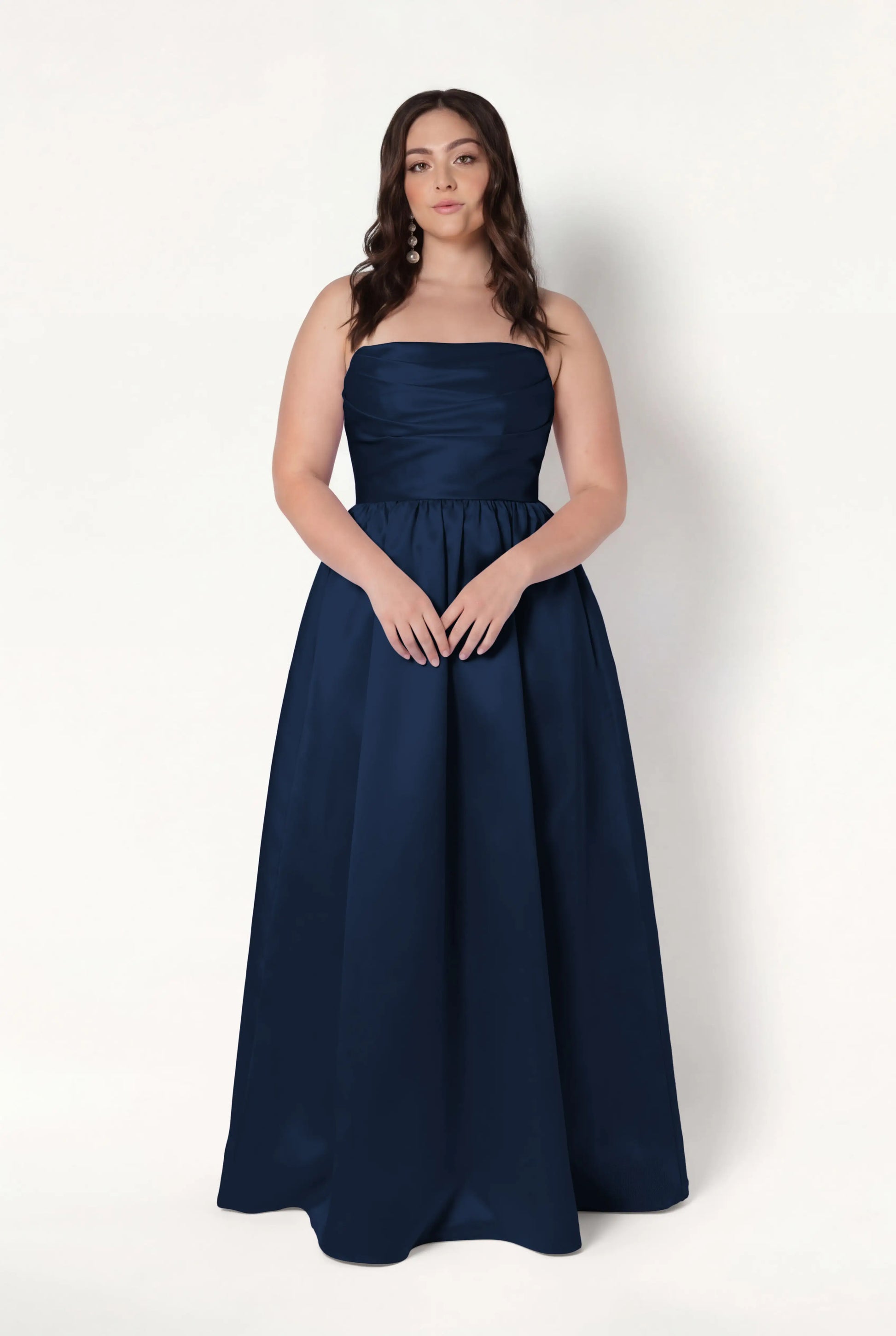 A woman with long brown hair wears the TH&TH Blair Bridesmaid Dress in Navy Satin Twill. She stands against a plain white background with her hands gently clasped in front of her.