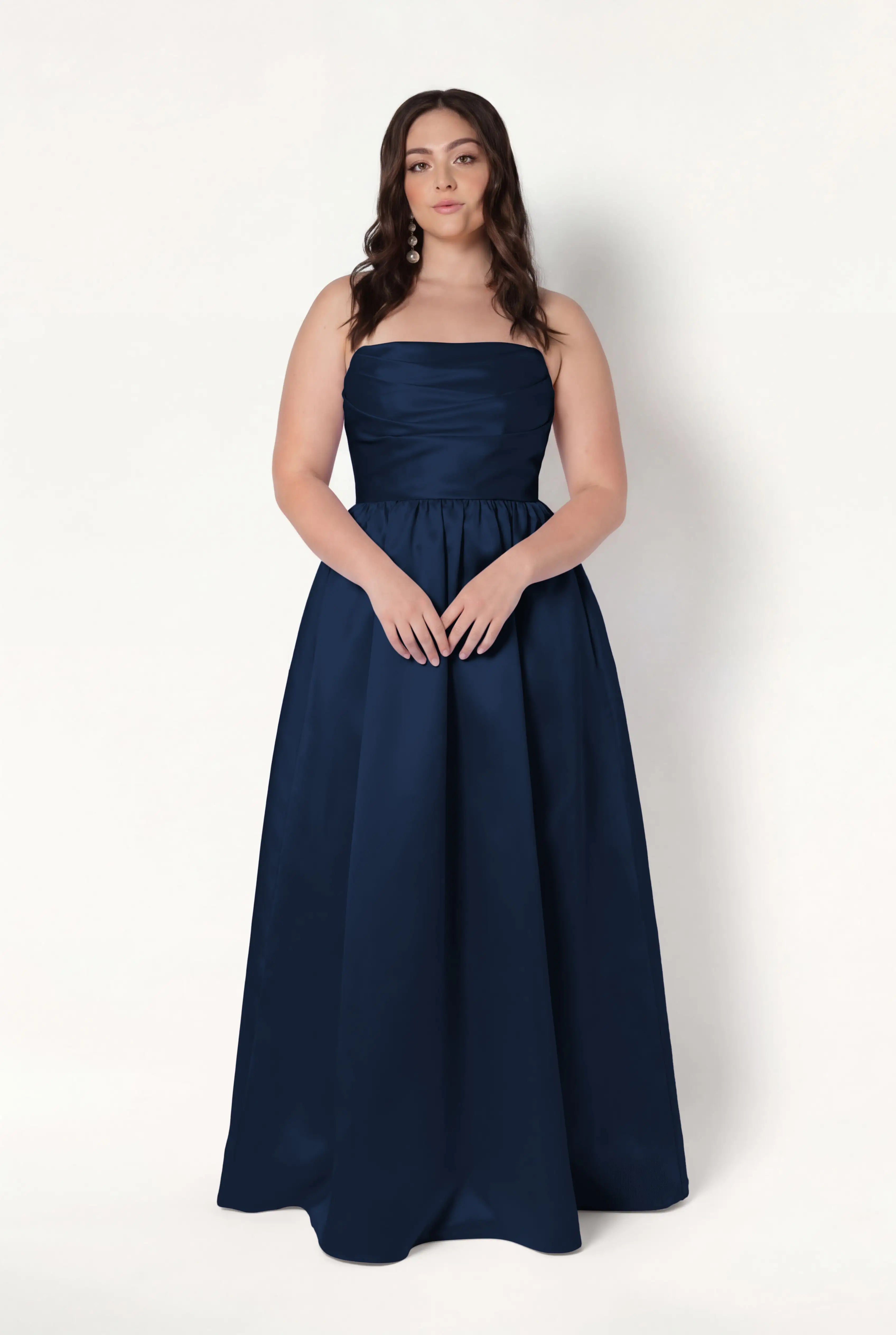 A woman with long brown hair wears the TH&TH Blair Bridesmaid Dress in Navy Satin Twill. She stands against a plain white background with her hands gently clasped in front of her.