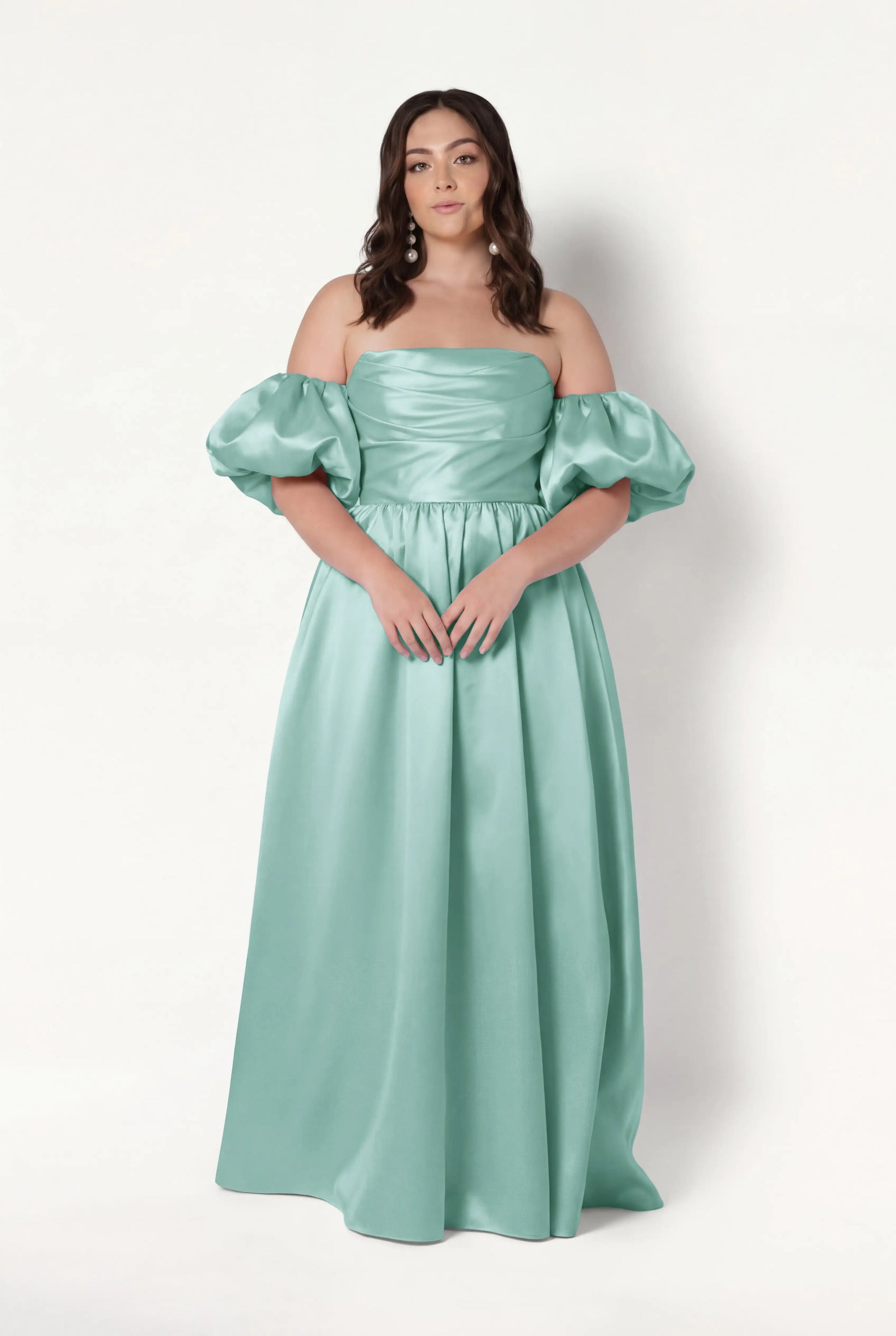 A woman poses against a white background in the TH&TH Blair Bridesmaid Dress in Seafoam Satin Twill, featuring a long off-the-shoulder corset gown with detachable puff sleeves and a fitted, ruched bodice. She has loose, wavy brown hair and a neutral expression.