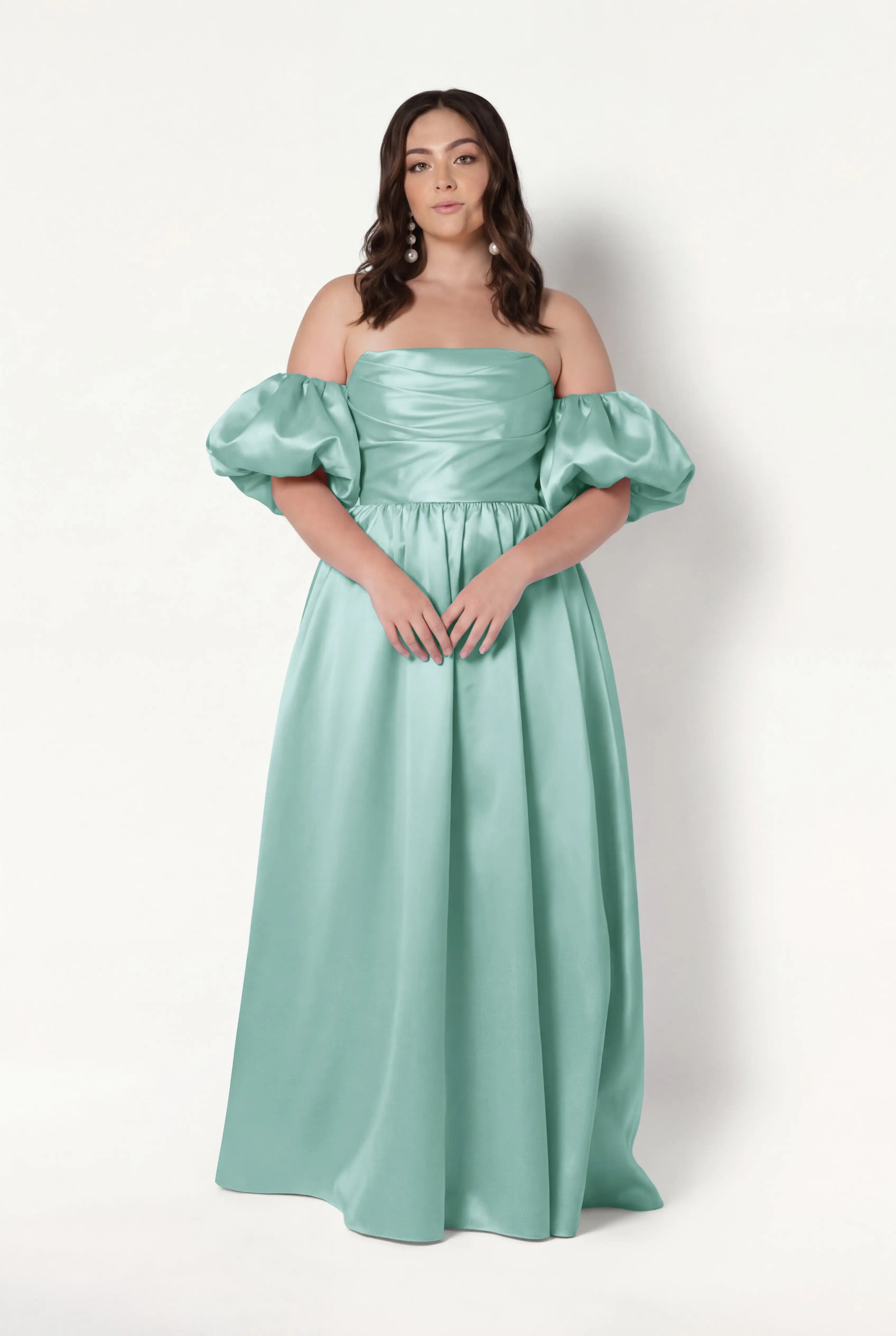 A woman poses against a white background in the TH&TH Blair Bridesmaid Dress in Seafoam Satin Twill, featuring a long off-the-shoulder corset gown with detachable puff sleeves and a fitted, ruched bodice. She has loose, wavy brown hair and a neutral expression.