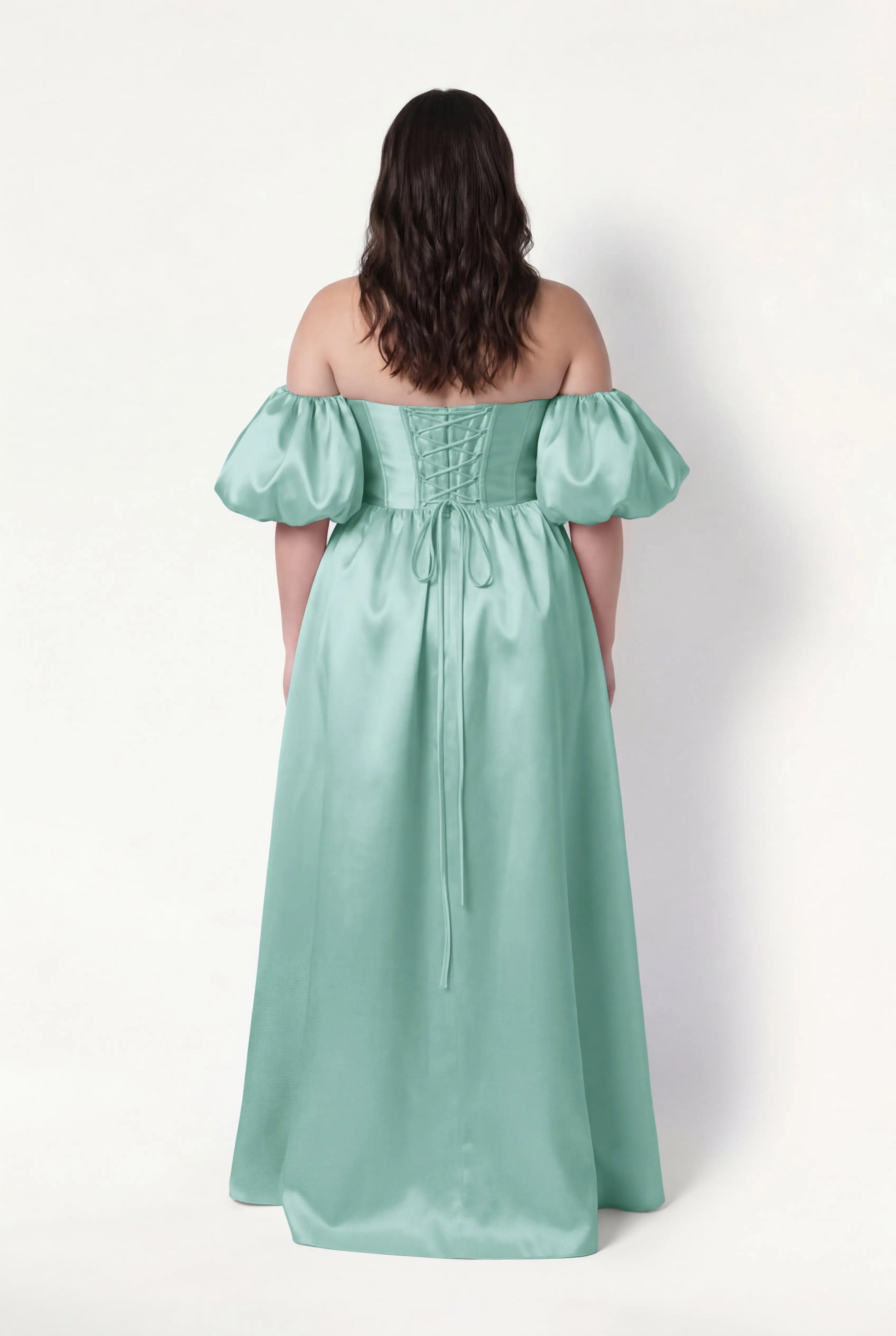 A woman with wavy dark hair stands facing away, wearing the TH&TH Blair Bridesmaid Dress in seafoam satin twill with off-the-shoulder, detachable puff sleeves and a lace-up back, set against a plain white background.