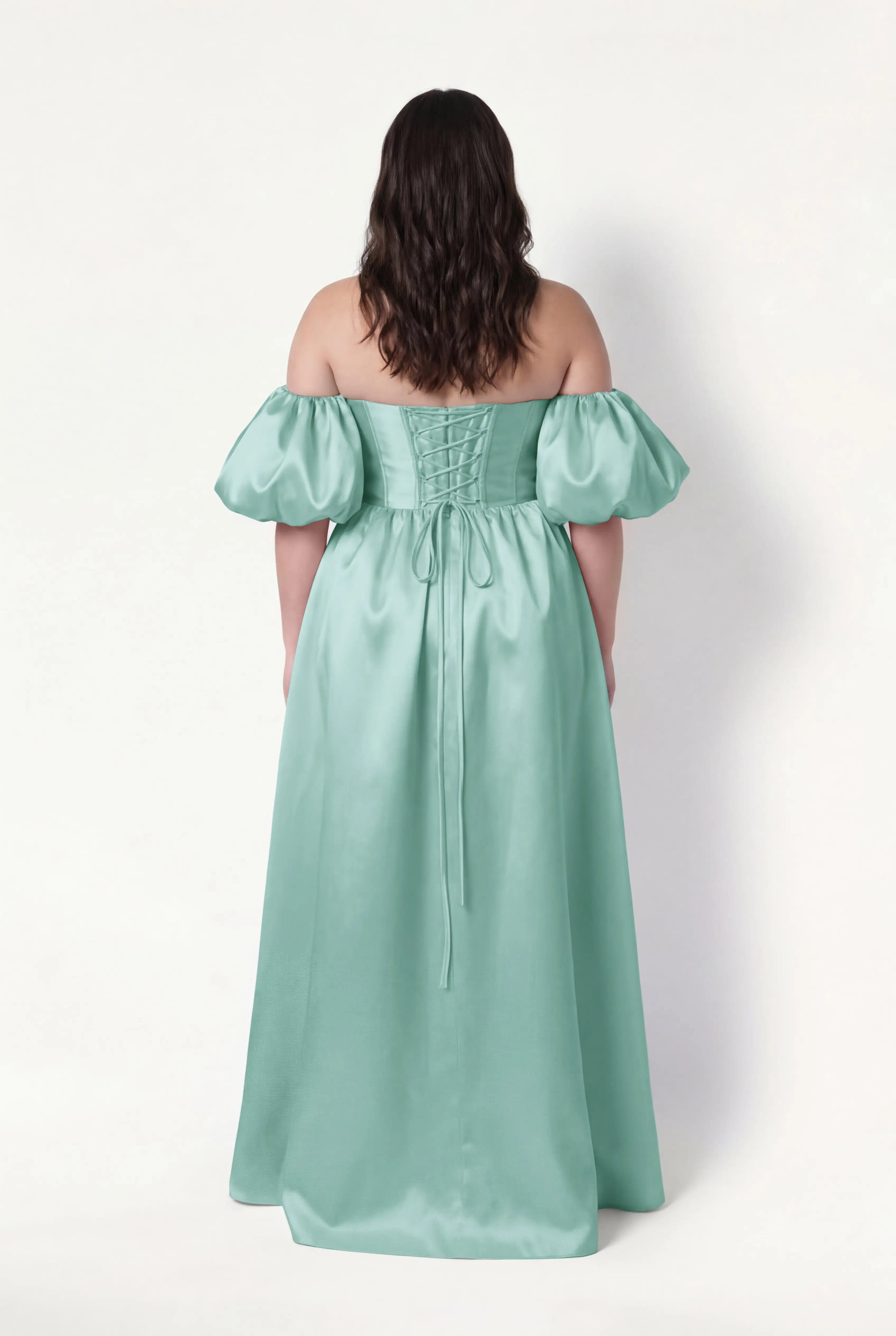 A woman with wavy dark hair stands facing away, wearing the TH&TH Blair Bridesmaid Dress in seafoam satin twill with off-the-shoulder, detachable puff sleeves and a lace-up back, set against a plain white background.