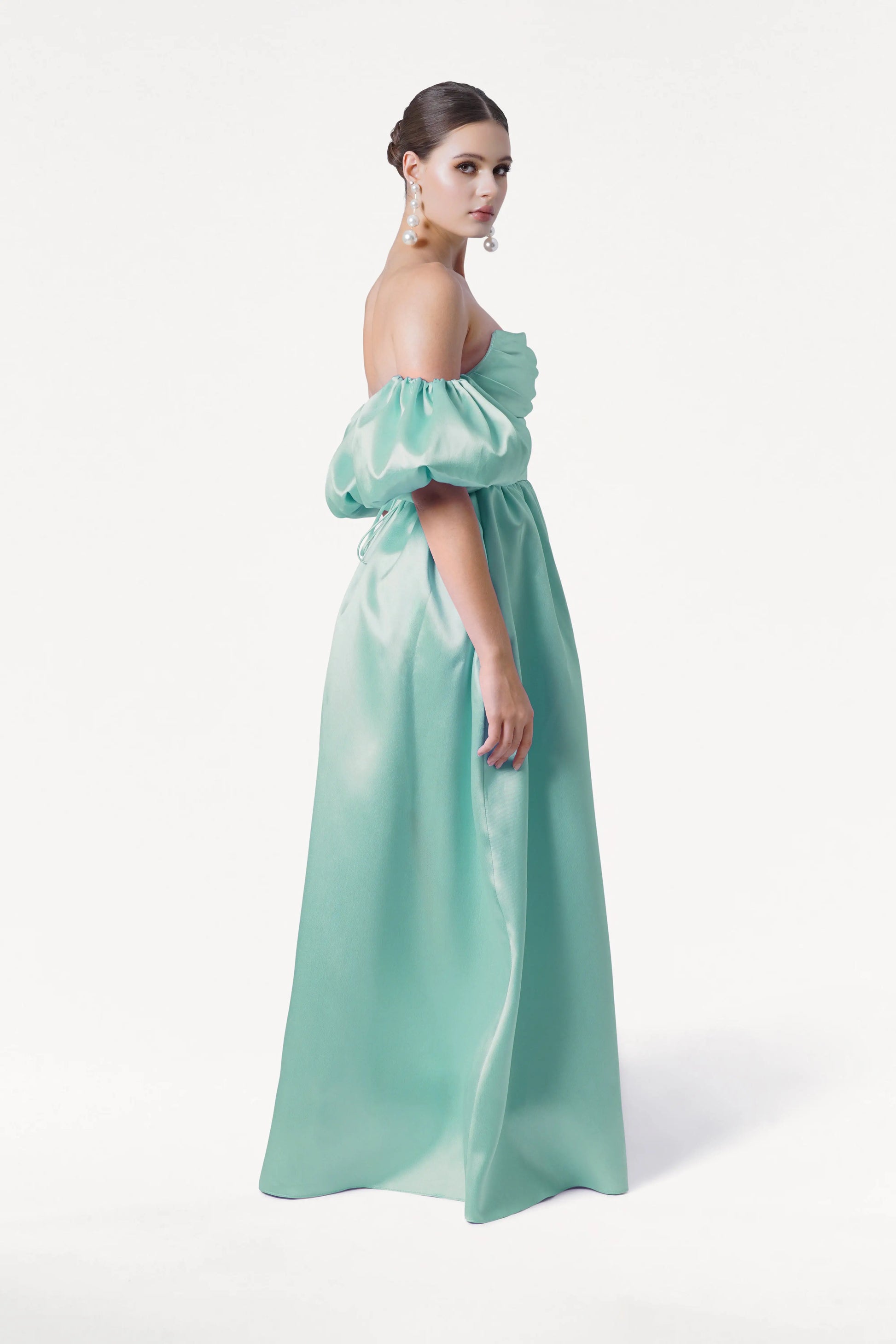 A woman poses sideways in the Blair Bridesmaid Dress in Seafoam Satin Twill by TH&TH, featuring detachable puff sleeves and a long skirt. She wears large pearl earrings, her hair in a sleek updo, against a plain white background.