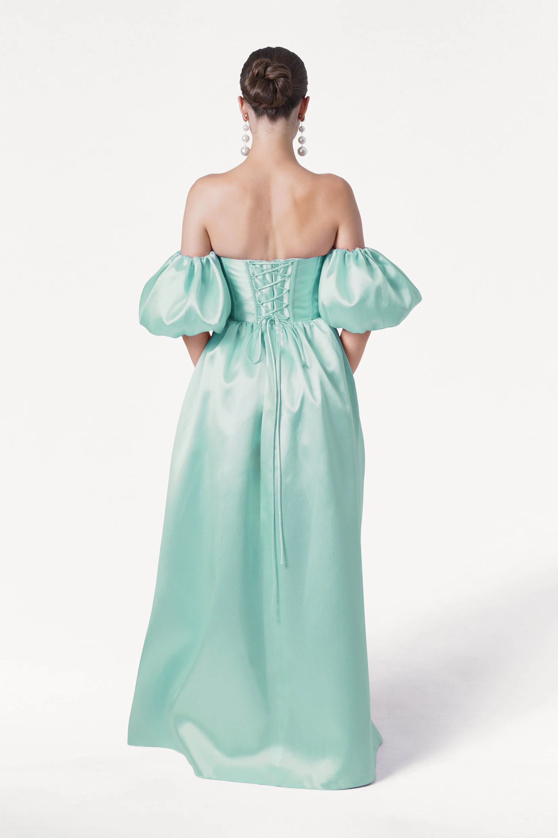 A woman faces away, wearing the TH&TH Blair Bridesmaid Dress in Seafoam Satin Twill with detachable puff sleeves and a lace-up back. Her hair is in a low bun, and she wears large dangling earrings against a plain white background.