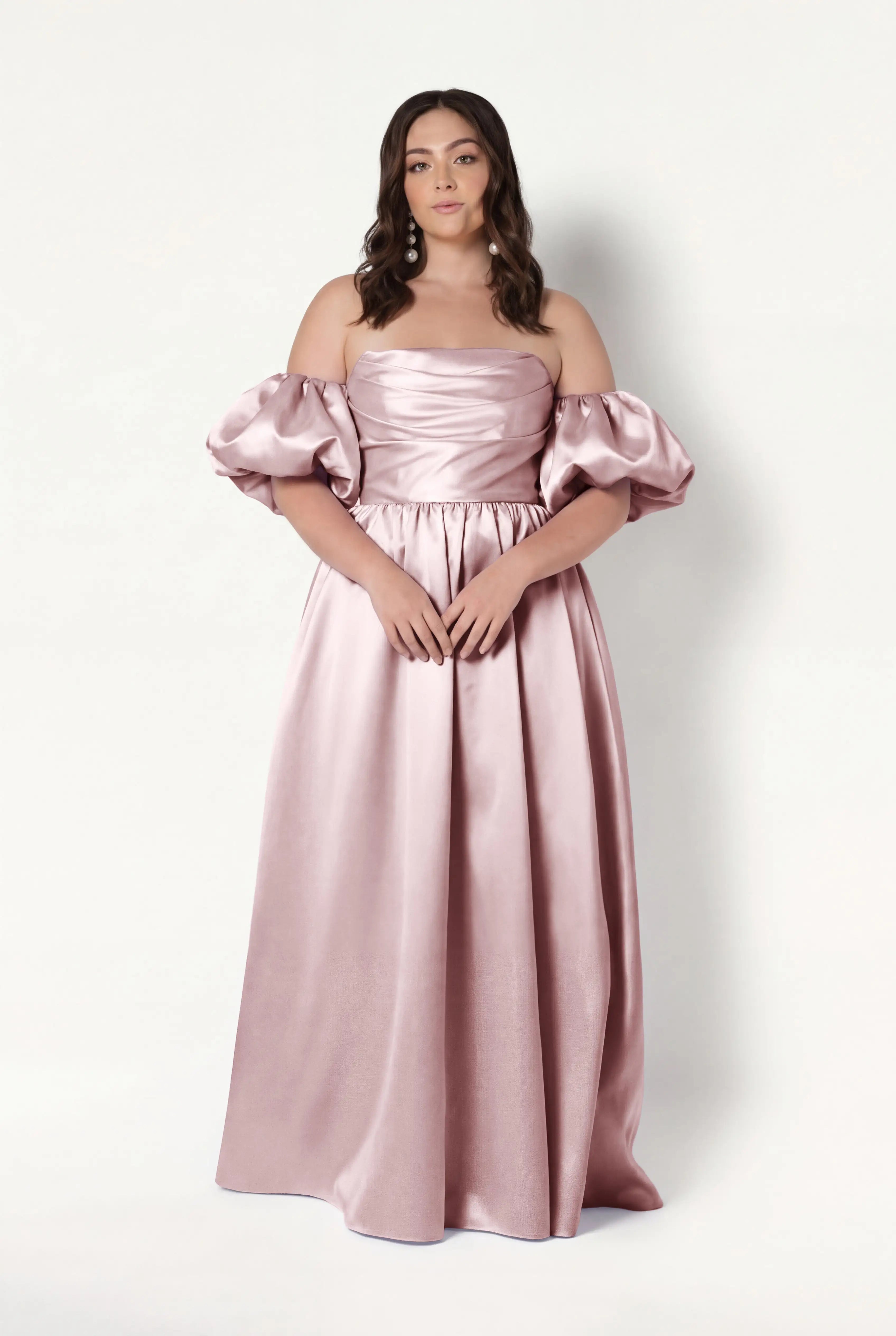A woman stands against a plain backdrop wearing the Blair Bridesmaid Dress in Smoked Blush Satin Twill by TH&TH—an off-the-shoulder, floor-length gown with detachable puff sleeves. She has long wavy brown hair and a calm expression.
