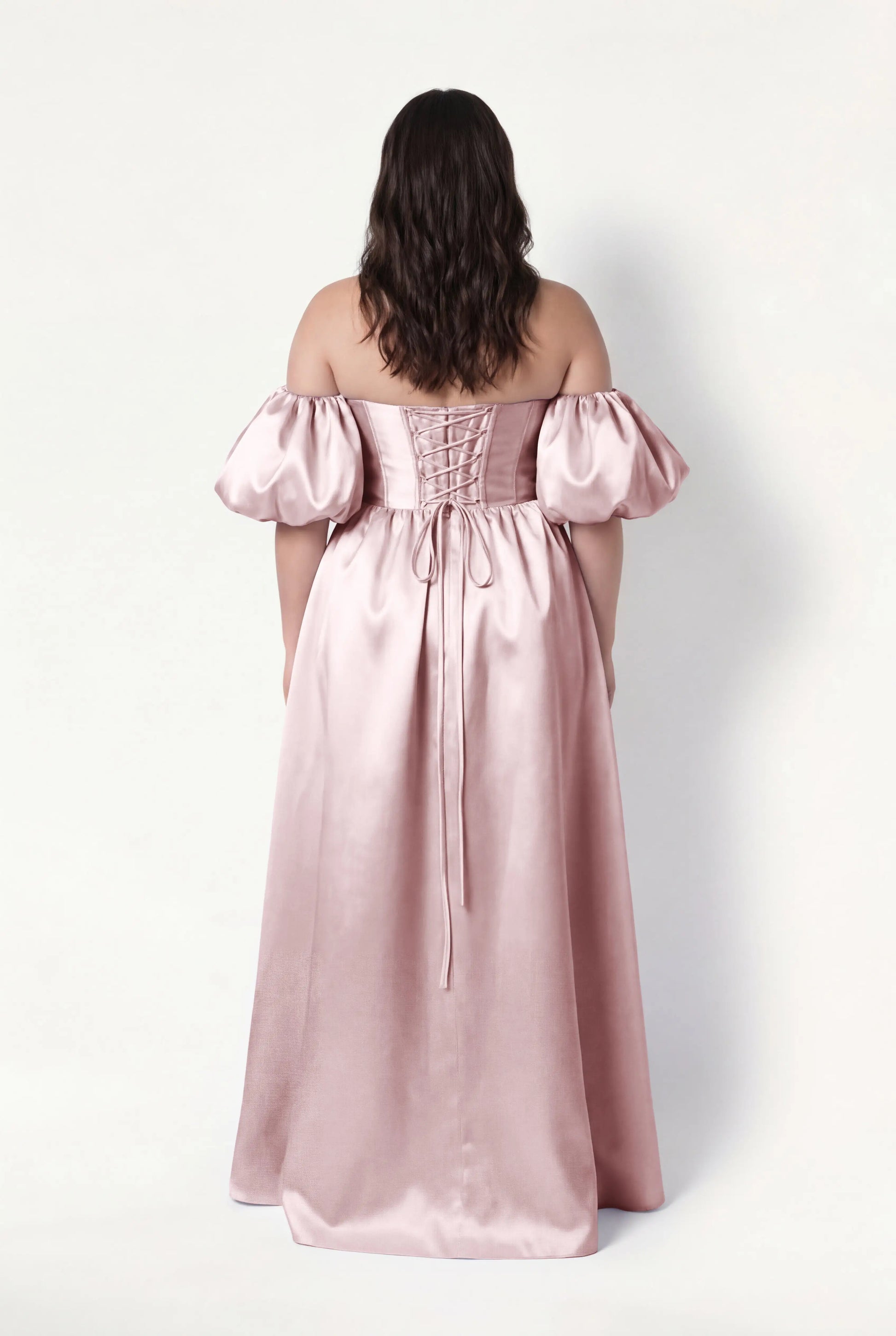 A woman with wavy brown hair faces away, wearing the TH&TH Blair Bridesmaid Dress in Smoked Blush Satin Twill with detachable puff sleeves and a lace-up back, standing against a plain white background.