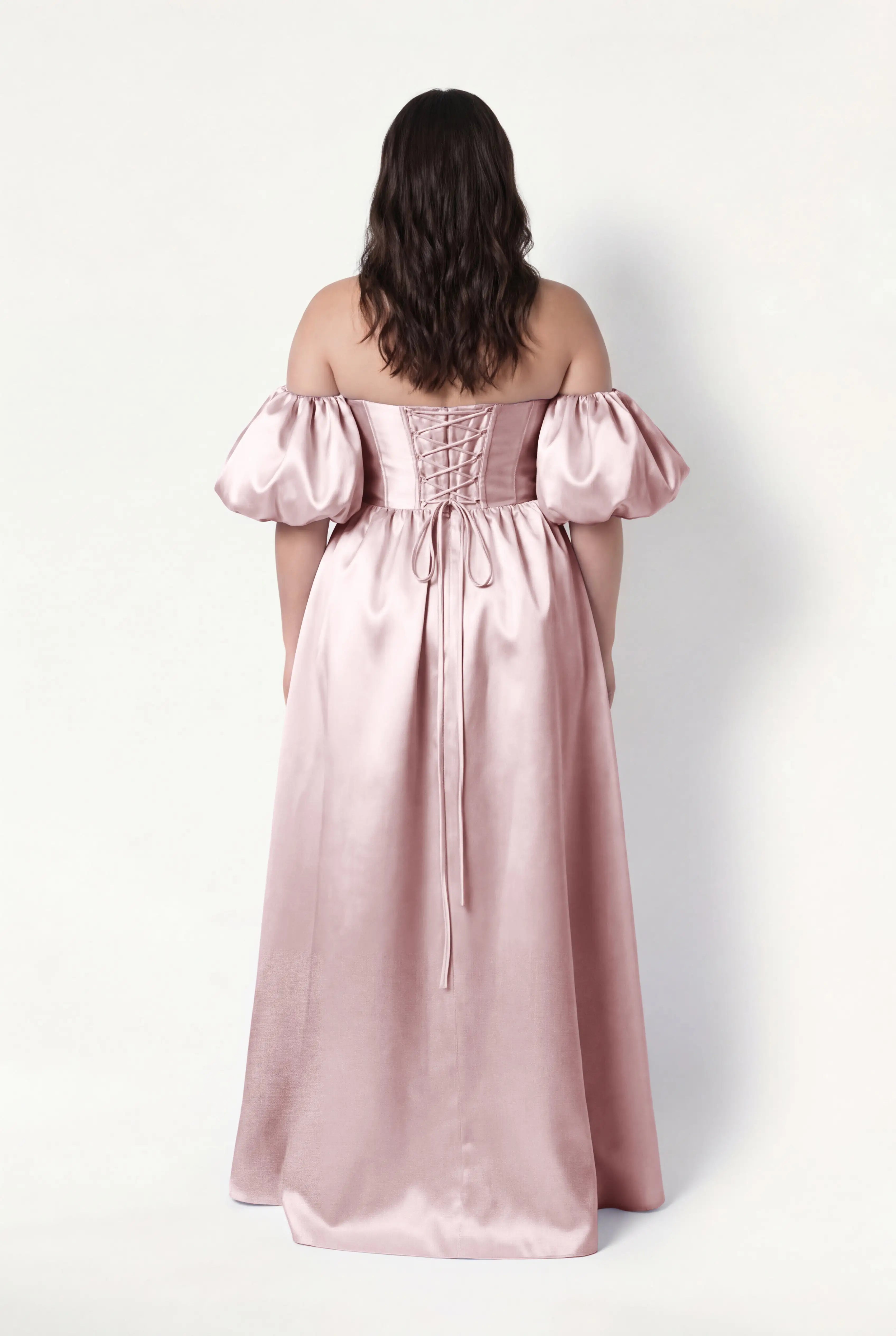 A woman with wavy brown hair faces away, wearing the TH&TH Blair Bridesmaid Dress in Smoked Blush Satin Twill with detachable puff sleeves and a lace-up back, standing against a plain white background.