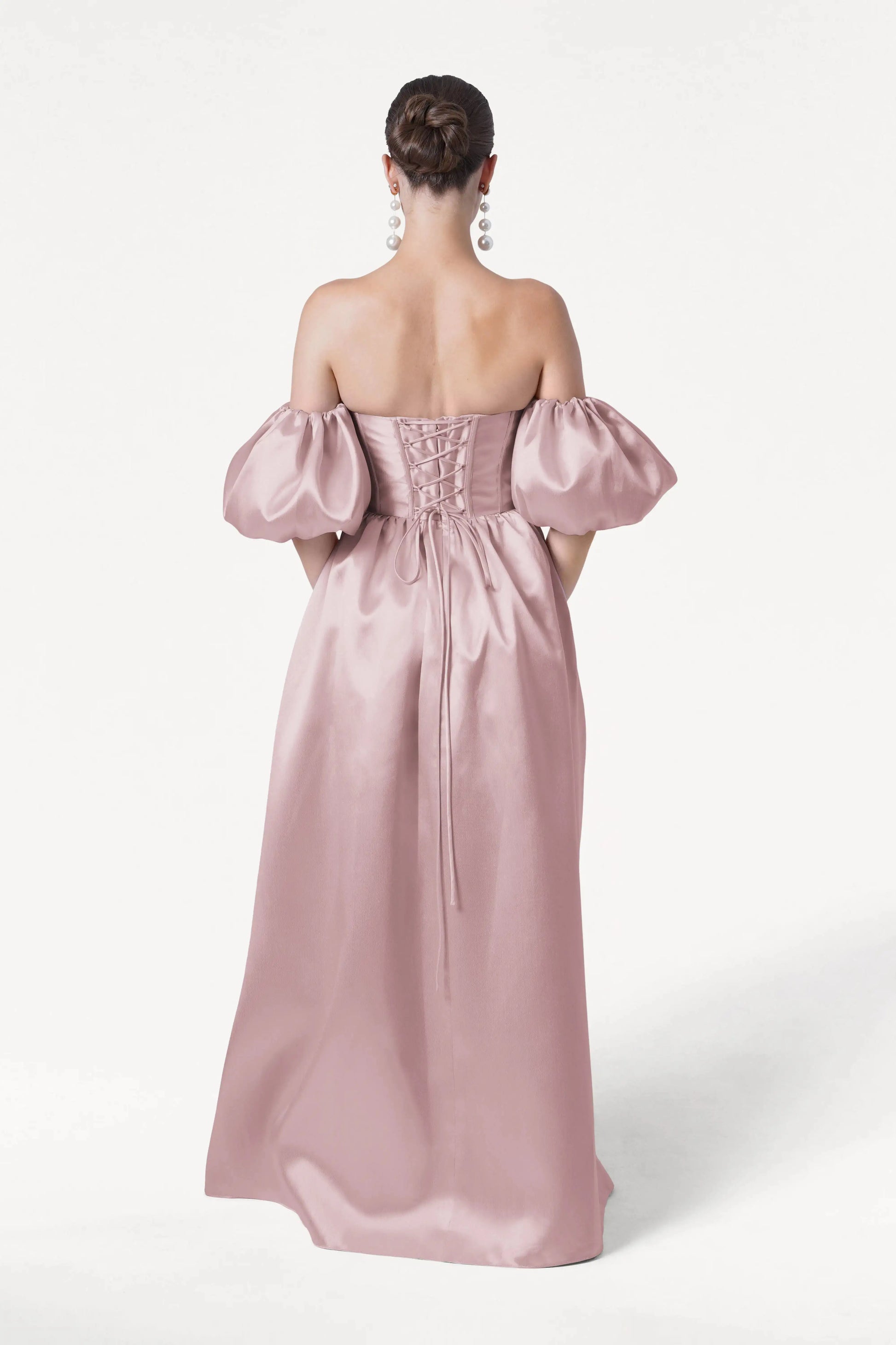 A woman stands facing away in the Blair Bridesmaid Dress by TH&TH, featuring smoked blush satin twill. Her hair is in a low bun, and she wears large dangling earrings, all set against a plain white background.
