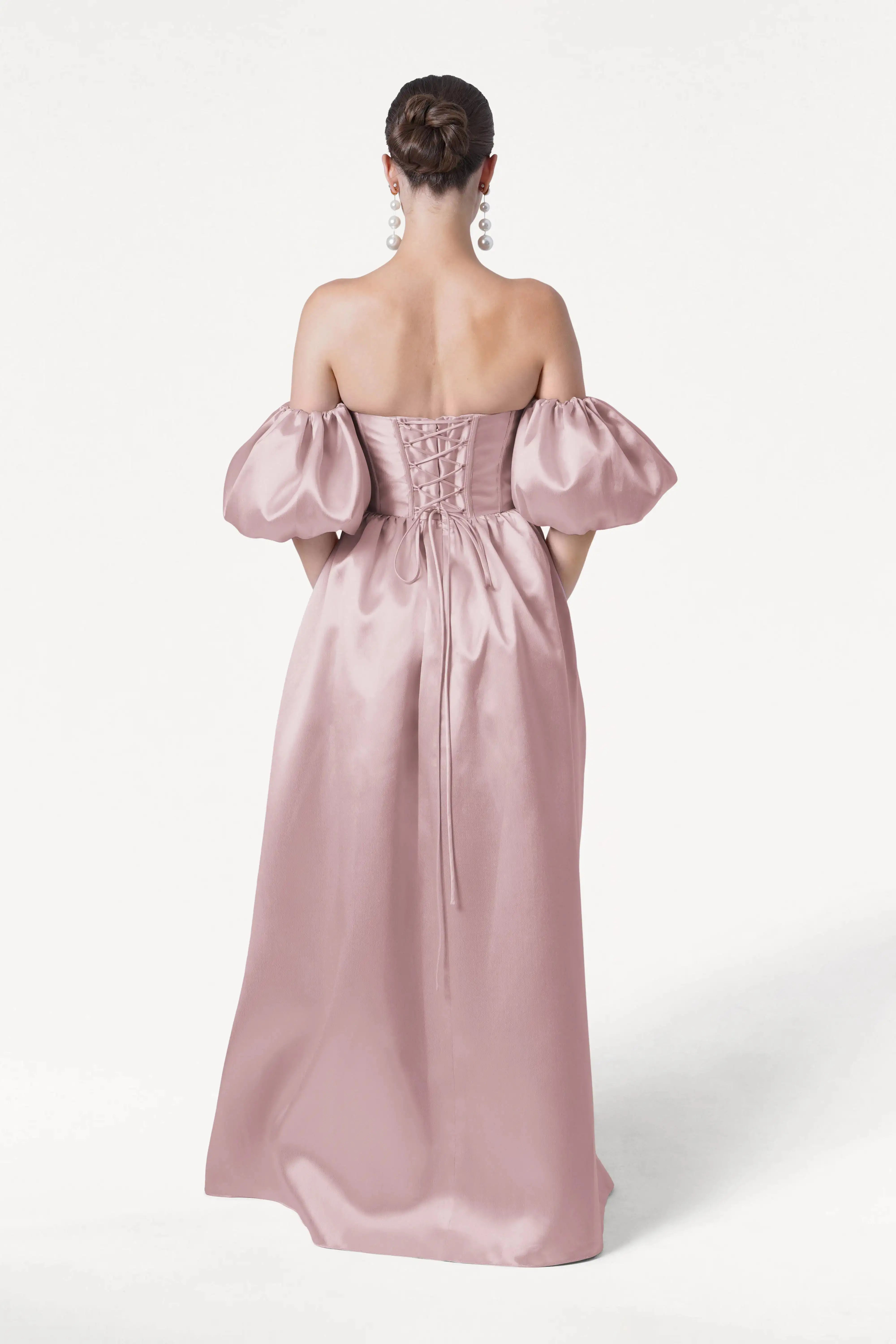 A woman stands facing away in the Blair Bridesmaid Dress by TH&TH, featuring smoked blush satin twill. Her hair is in a low bun, and she wears large dangling earrings, all set against a plain white background.