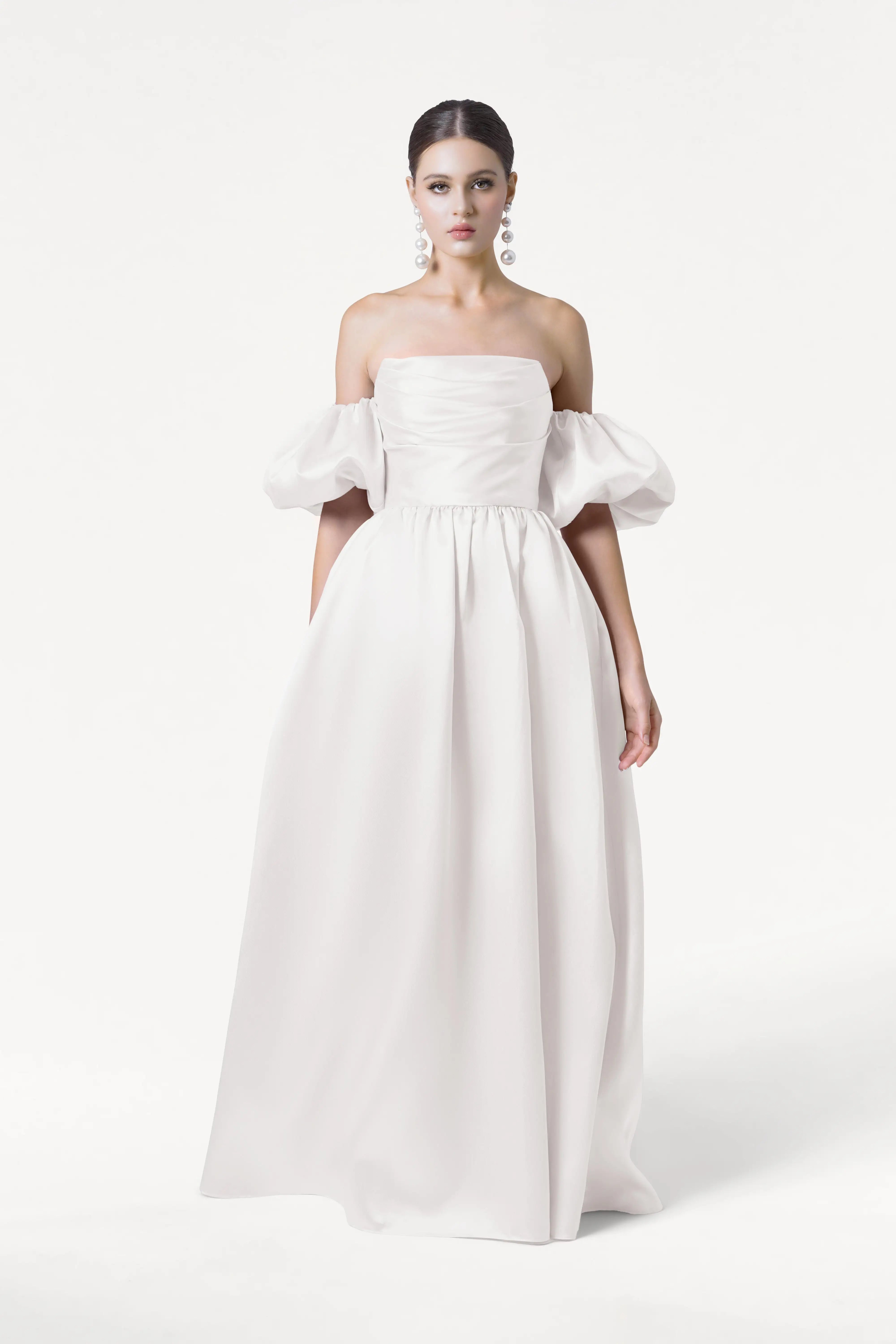 A woman poses against a plain white background in the TH&TH Blair Dress in Ivory Satin Twill, featuring an off-the-shoulder ruched bodice, detachable puff sleeves, and a floor-length skirt. Her dark hair is pulled back, accessorized by large dangling earrings.