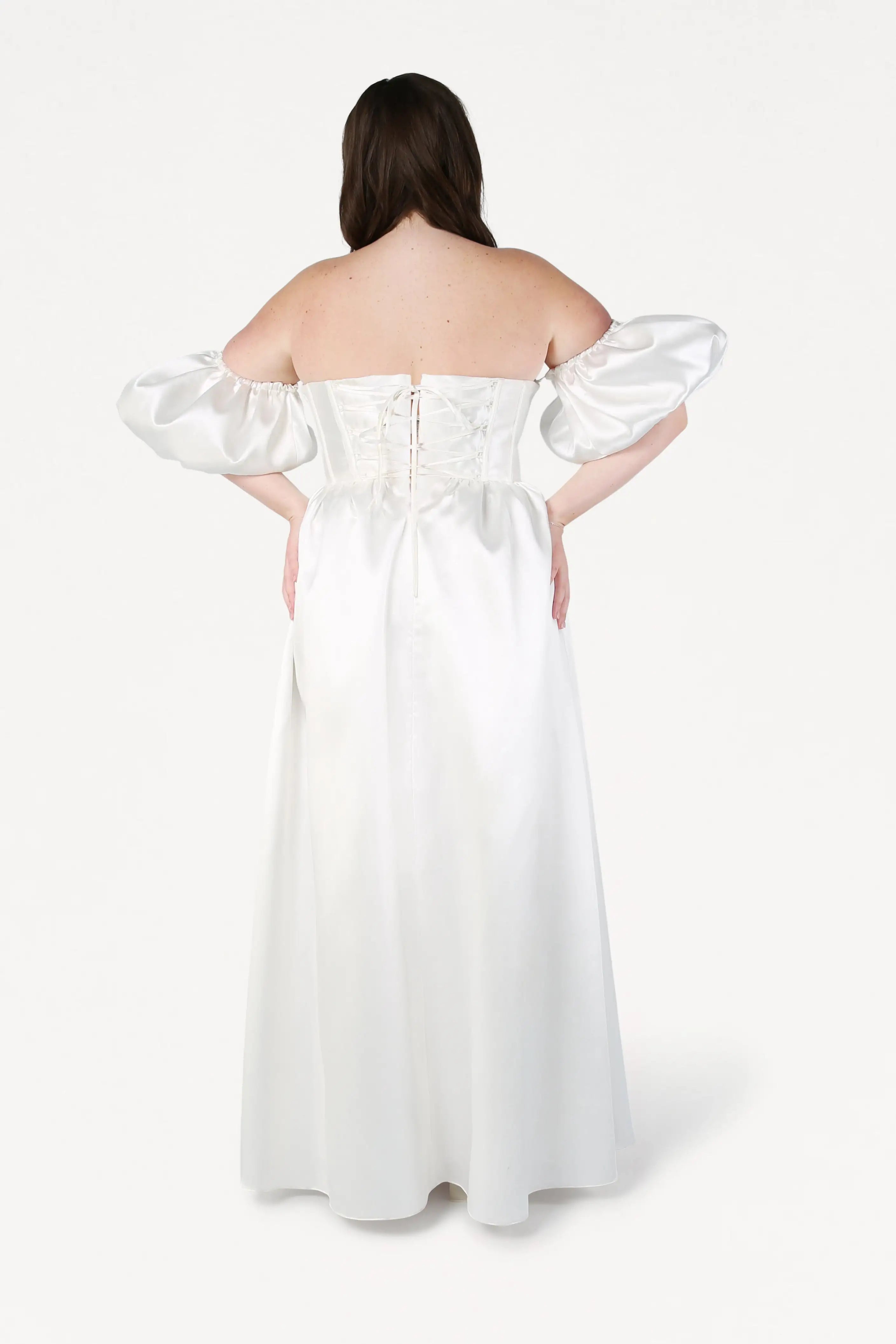 A woman faces away from the camera in the TH&TH Blair Dress in Ivory Satin Twill, with detachable puff sleeves, a ruched bodice, floor-length skirt, satin sheen, and a lace-up back.