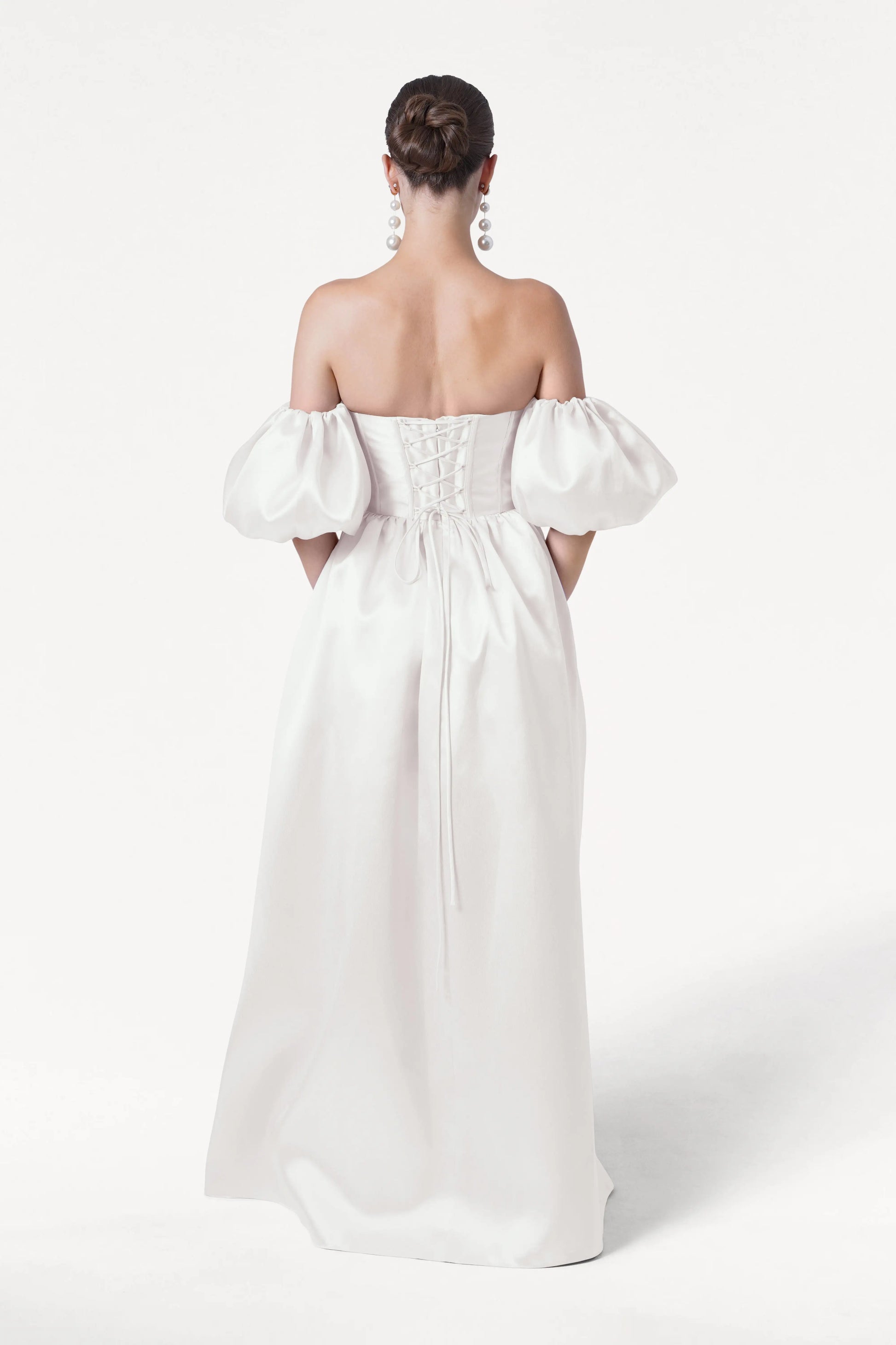 A woman with brown hair in a low bun wears the TH&TH Blair Dress in Ivory Satin Twill, featuring an off-the-shoulder neckline, detachable puff sleeves, and a lace-up back. She faces away from the camera against a plain white background.