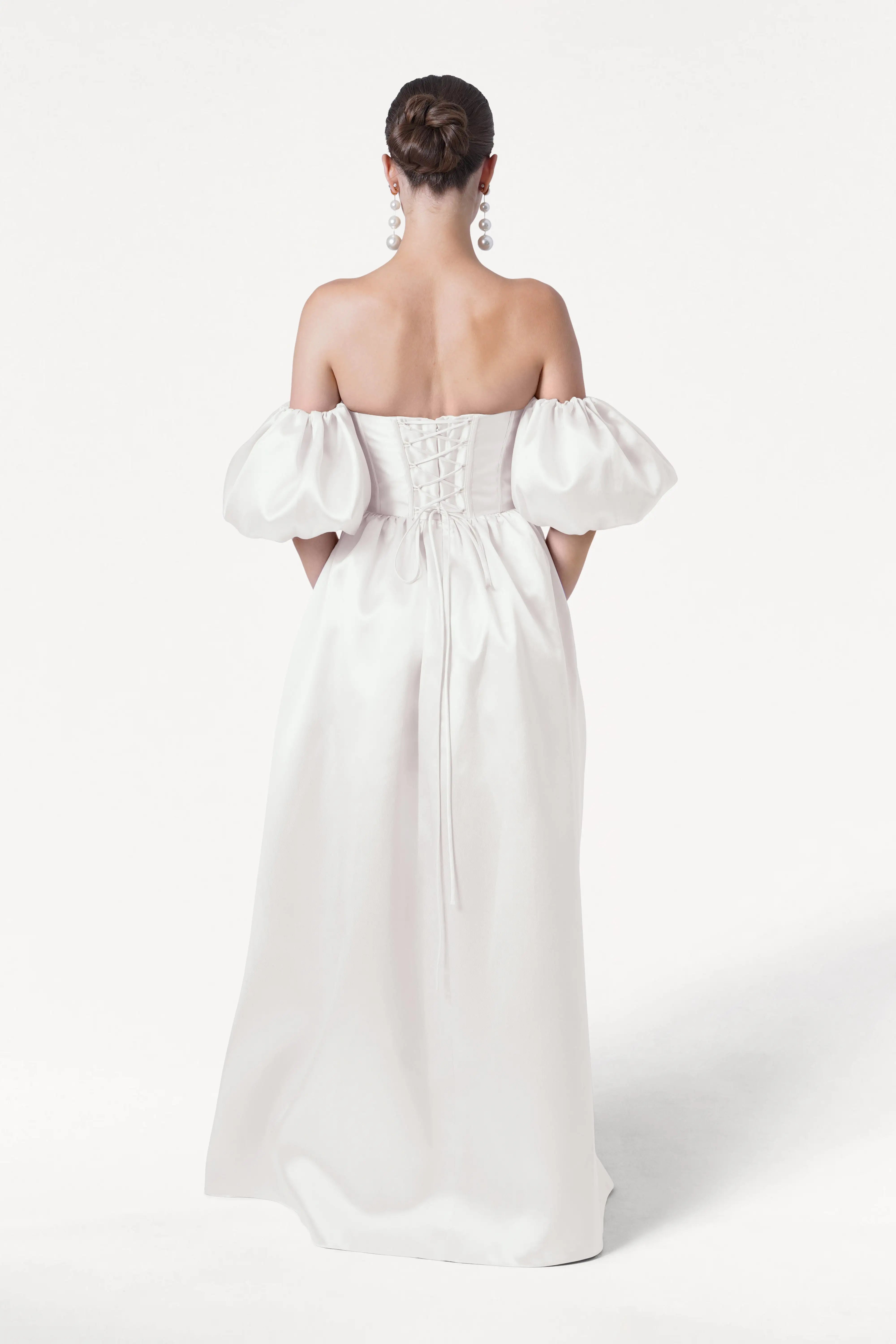 A woman with brown hair in a low bun wears the TH&TH Blair Dress in Ivory Satin Twill, featuring an off-the-shoulder neckline, detachable puff sleeves, and a lace-up back. She faces away from the camera against a plain white background.