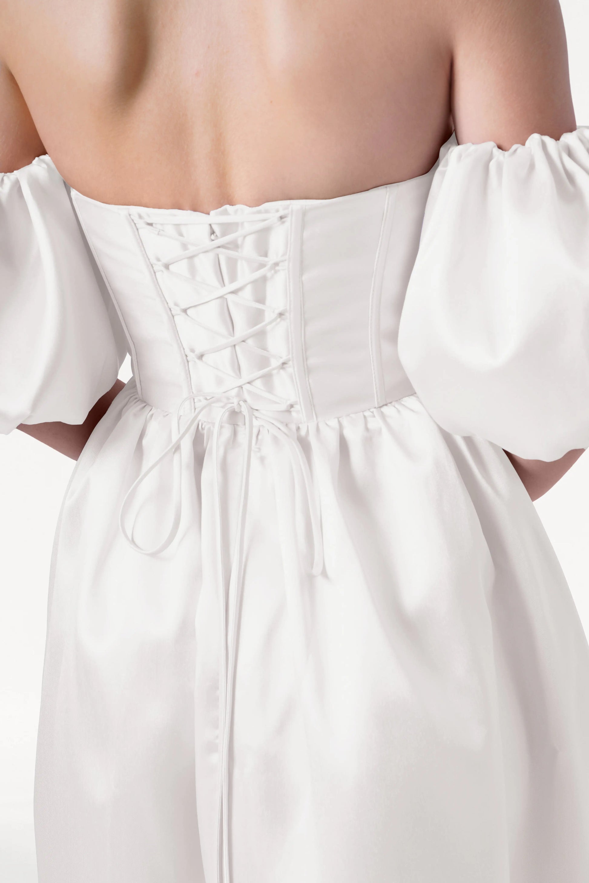 A close-up shows someone wearing the TH&TH Blair Dress in Ivory Satin Twill, an off-the-shoulder wedding gown with smooth, shiny fabric, detachable puff sleeves, and a lace-up back detail photographed from behind on a plain background.