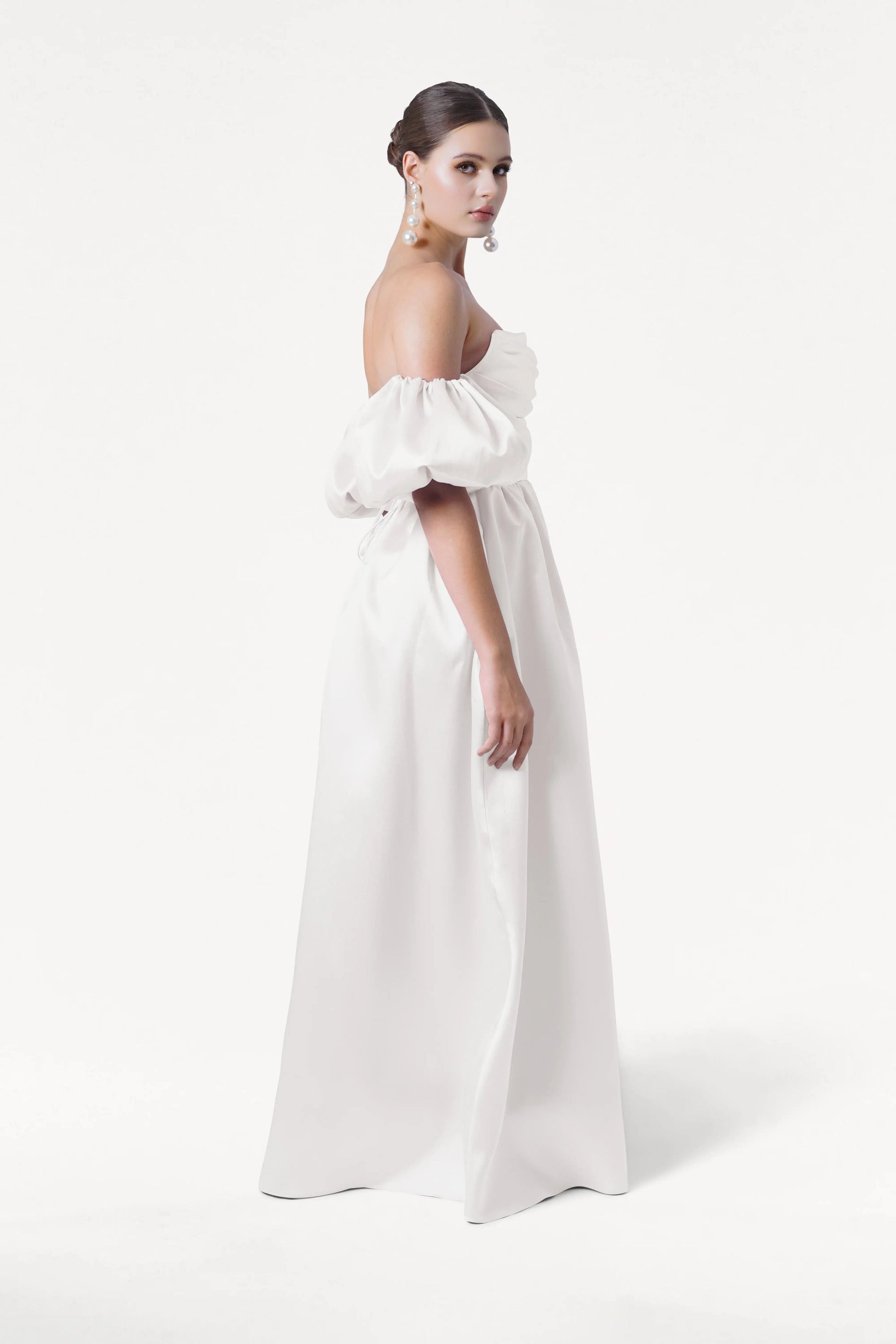 A woman wears the Blair Dress in Ivory Satin Twill by TH&TH, featuring a white off-shoulder ruched bodice and detachable puff sleeves. She stands against a white background, looking over her shoulder with an elegant updo and statement earrings.