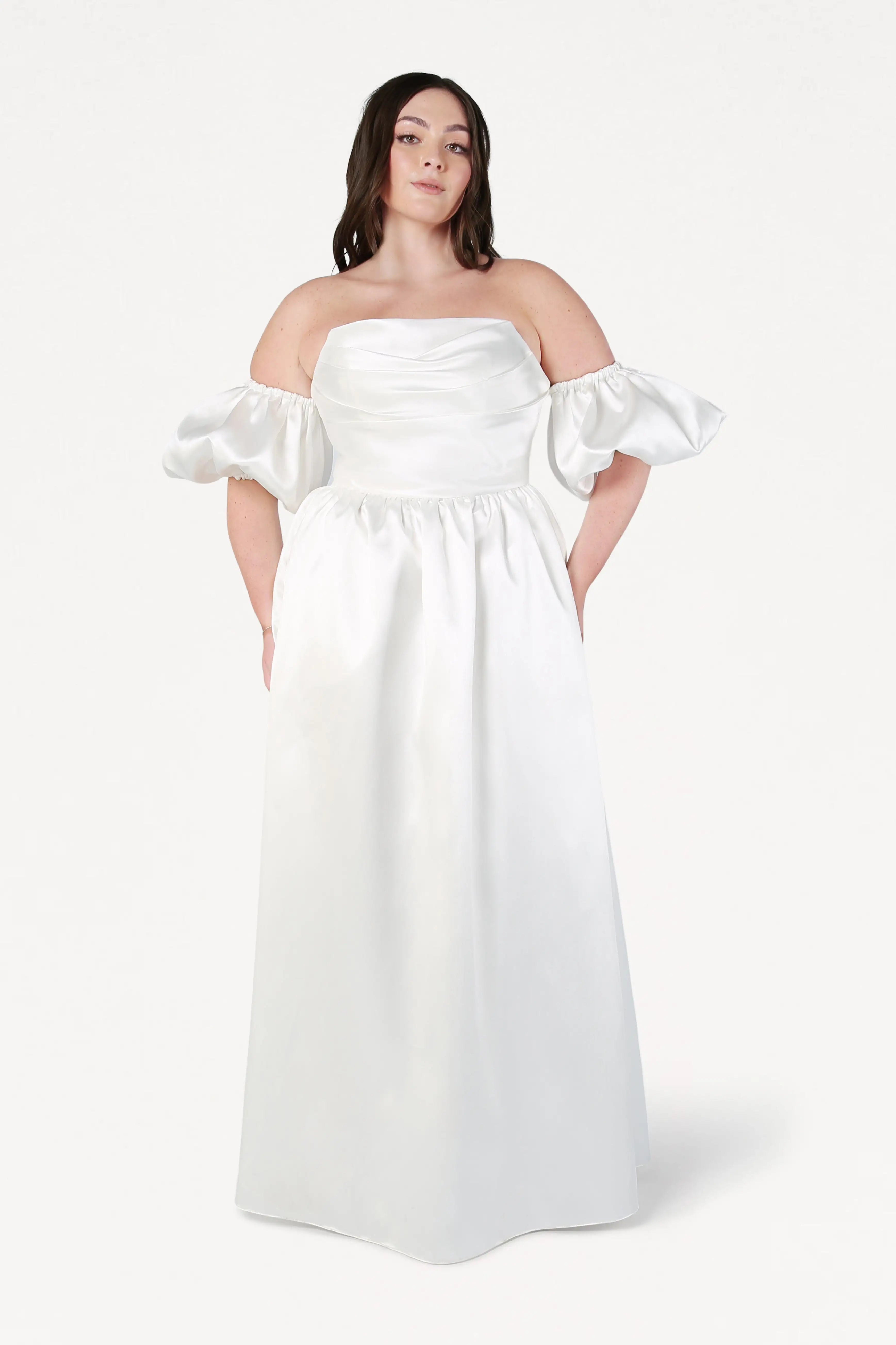 A woman wearing the Blair Dress in Ivory Satin Twill by TH&TH, a white off-the-shoulder, floor-length corset wedding gown with detachable puff sleeves, stands against a plain white background.