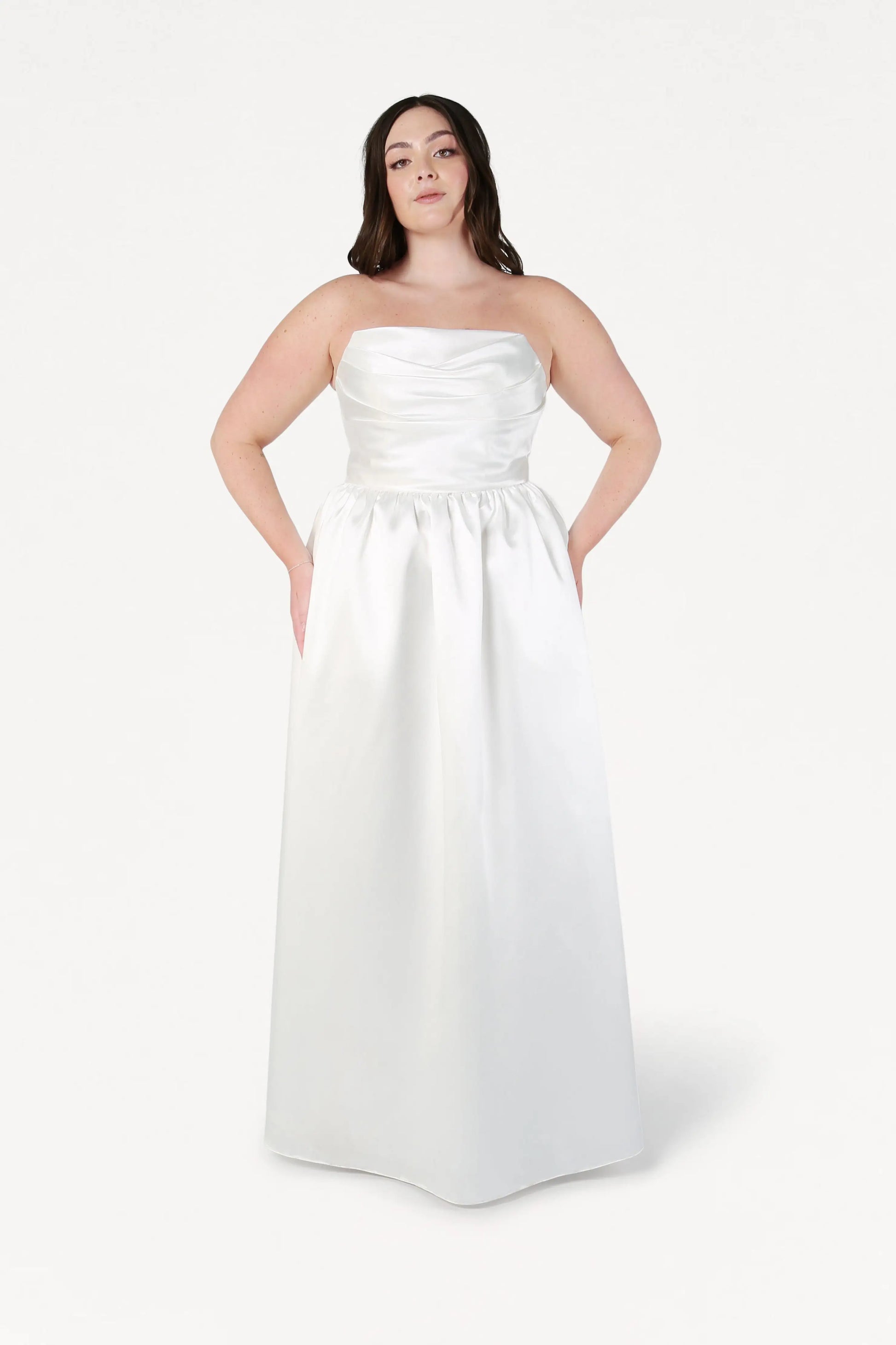 A woman wearing the Blair Dress in Ivory Satin Twill by TH&TH, a strapless floor-length gown with a ruched bodice and pockets, stands facing forward against a plain white background.