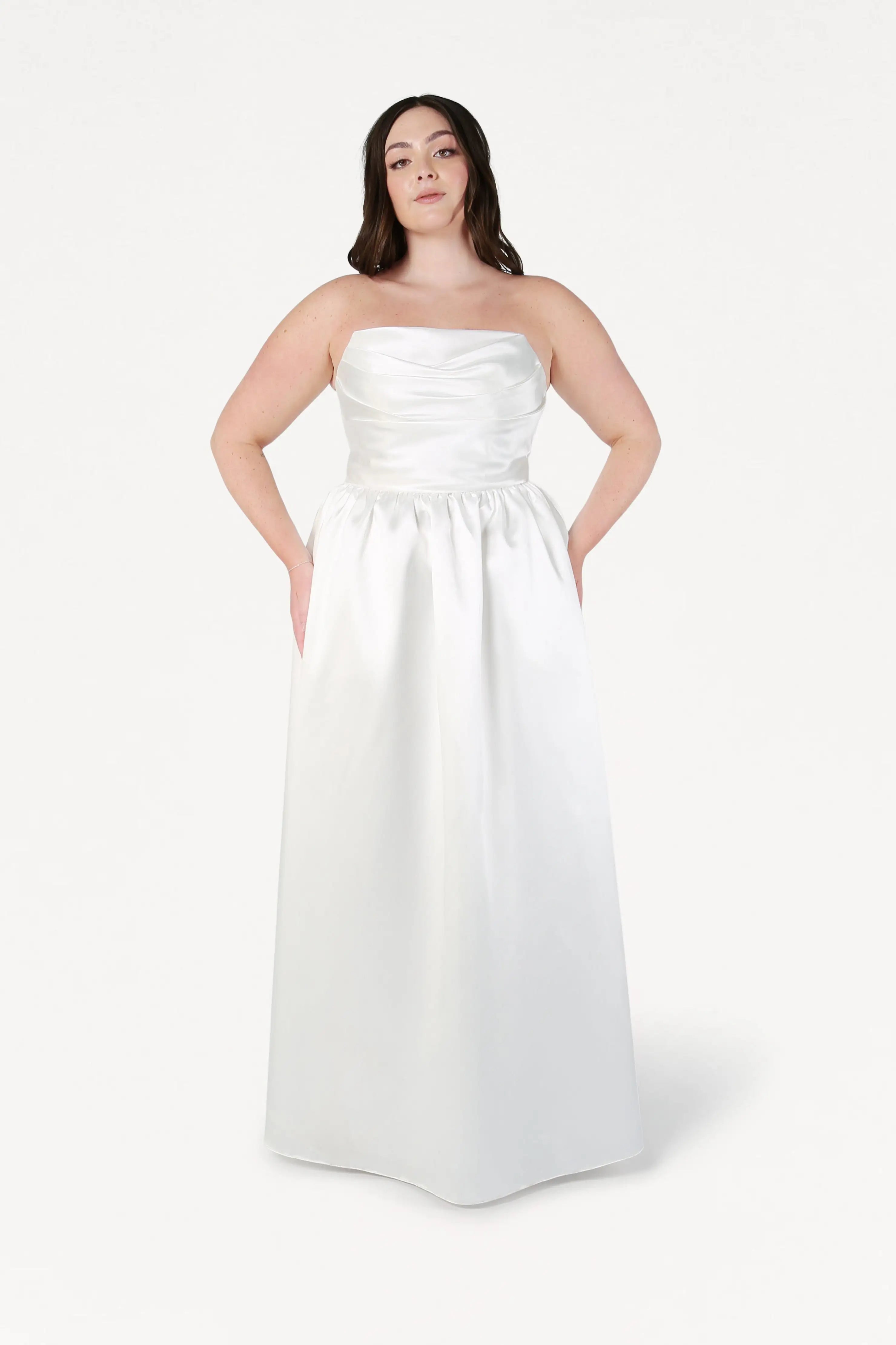 A woman wearing the Blair Dress in Ivory Satin Twill by TH&TH, a strapless floor-length gown with a ruched bodice and pockets, stands facing forward against a plain white background.