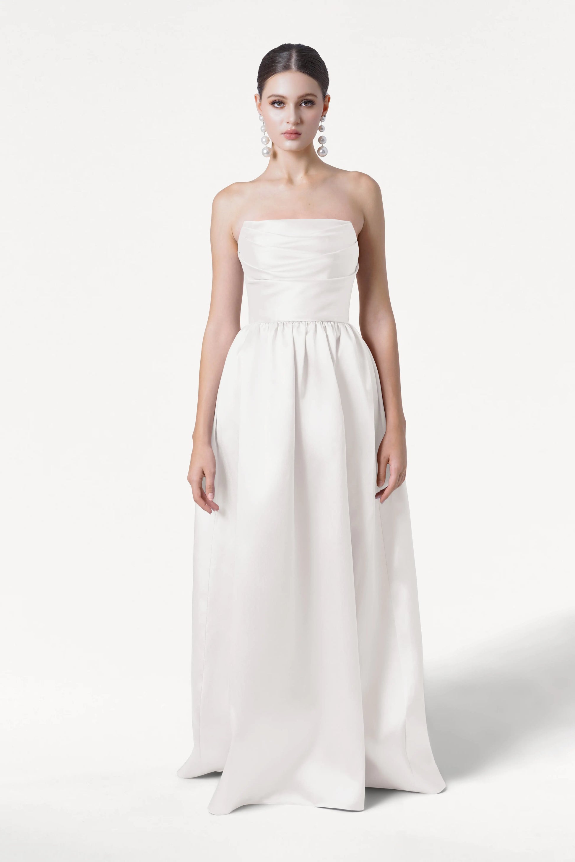 A woman poses against a white background in the Blair Dress in Ivory Satin Twill by TH&TH. The strapless, floor-length gown features a ruched fitted bodice, full skirt, detachable puff sleeves, and large pearl earrings with her hair pulled back.