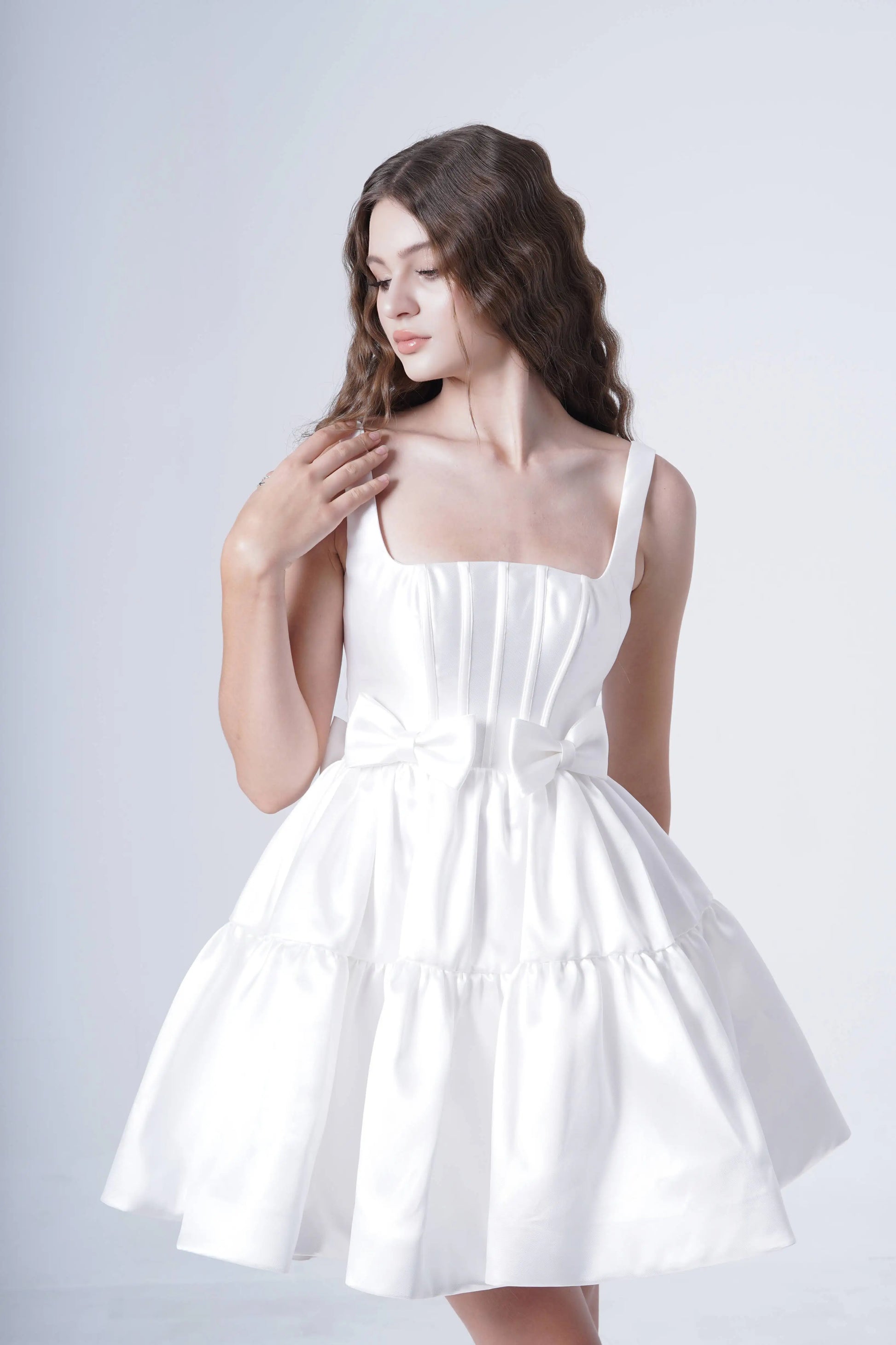 A woman wearing the TH&TH Bloom Bridal Mini Dress in Ivory, a sleeveless white satin twill mini with a waist bow, stands against a plain light background, looking down and touching her wavy hair.