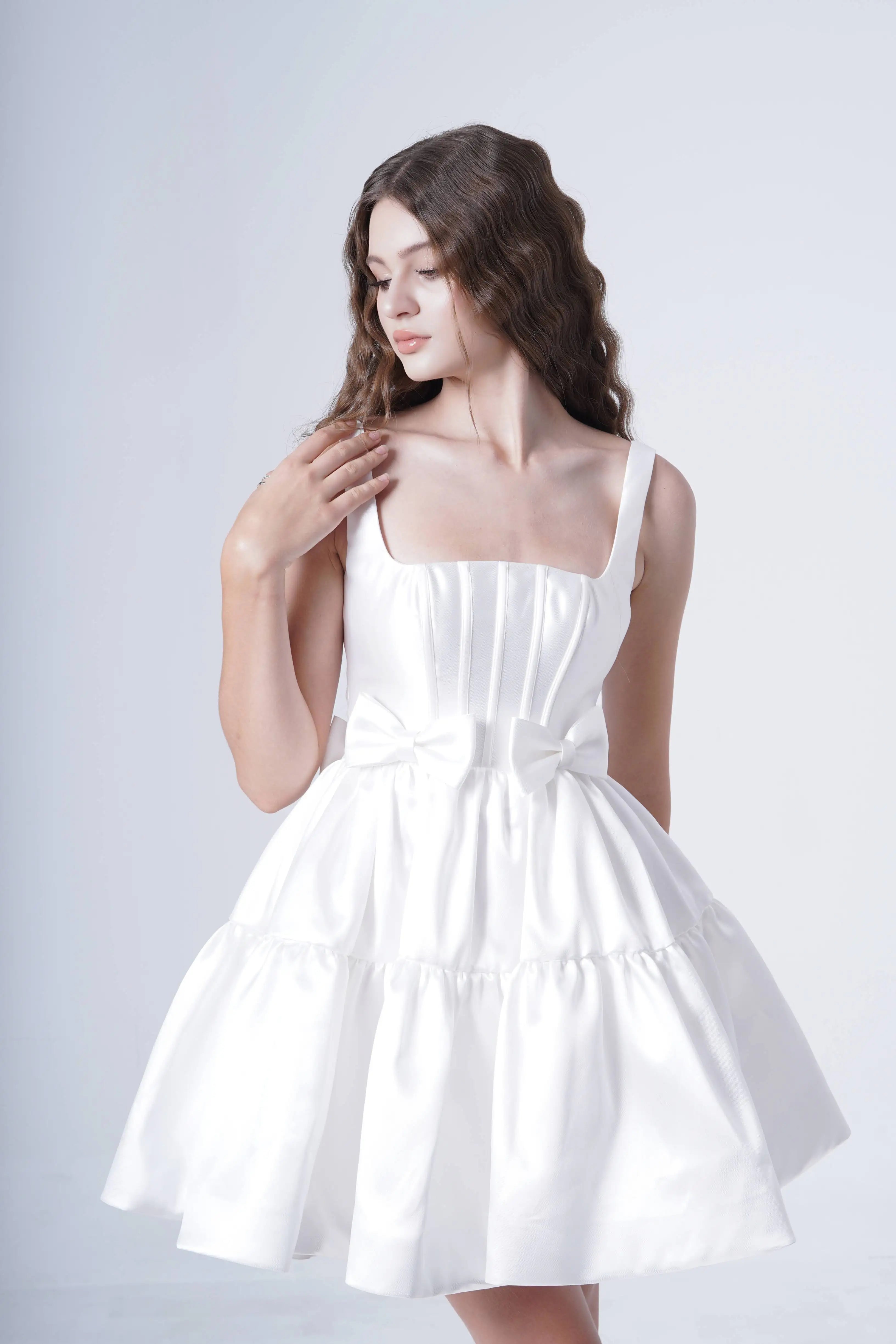 A woman wearing the TH&TH Bloom Bridal Mini Dress in Ivory, a sleeveless white satin twill mini with a waist bow, stands against a plain light background, looking down and touching her wavy hair.