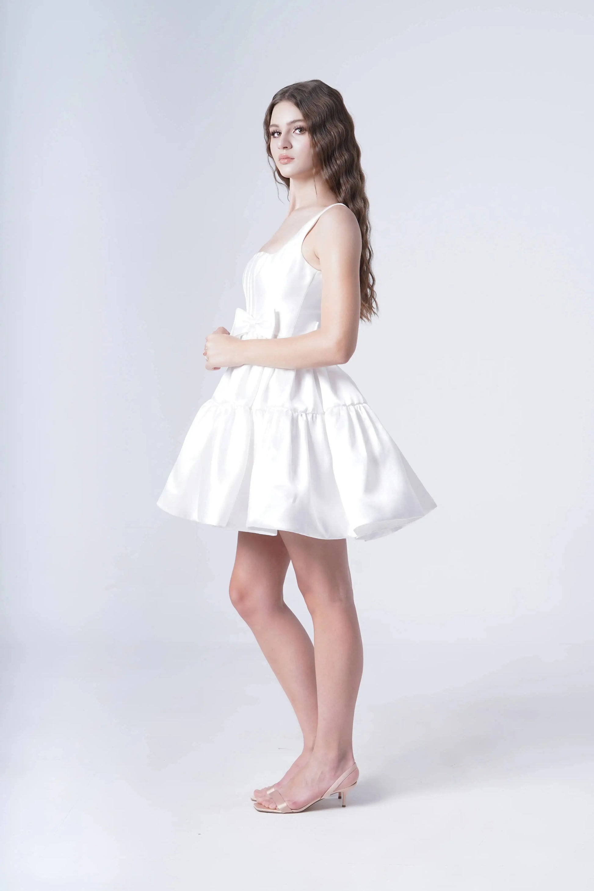 A young woman with long wavy hair models the TH&TH Bloom Bridal Mini Dress in Ivory, featuring a corseted bodice and sleeveless mini cut, paired with clear heels as she poses sideways against a plain white background.