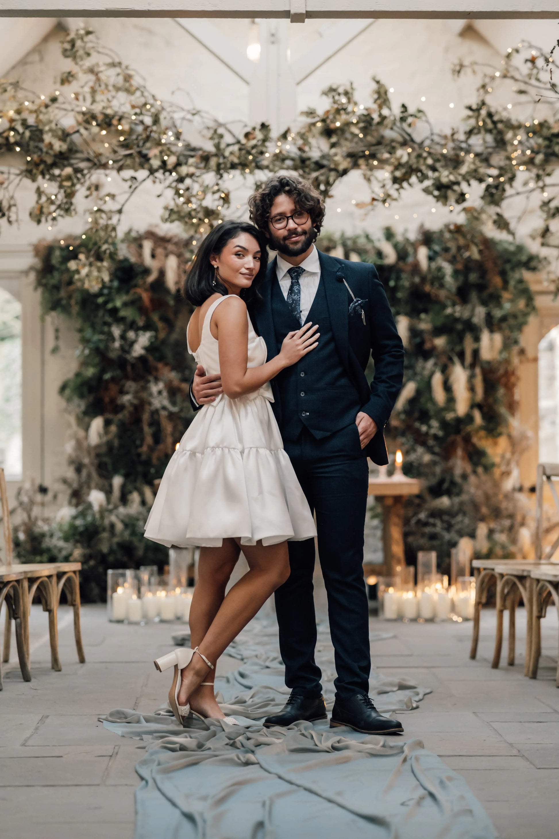 A couple poses at a wedding; she wears the TH&TH Bloom Bridal Mini Dress in Ivory with heels, while he is in a dark suit and tie. They stand on a candle-lit, greenery-lined aisle surrounded by chairs.