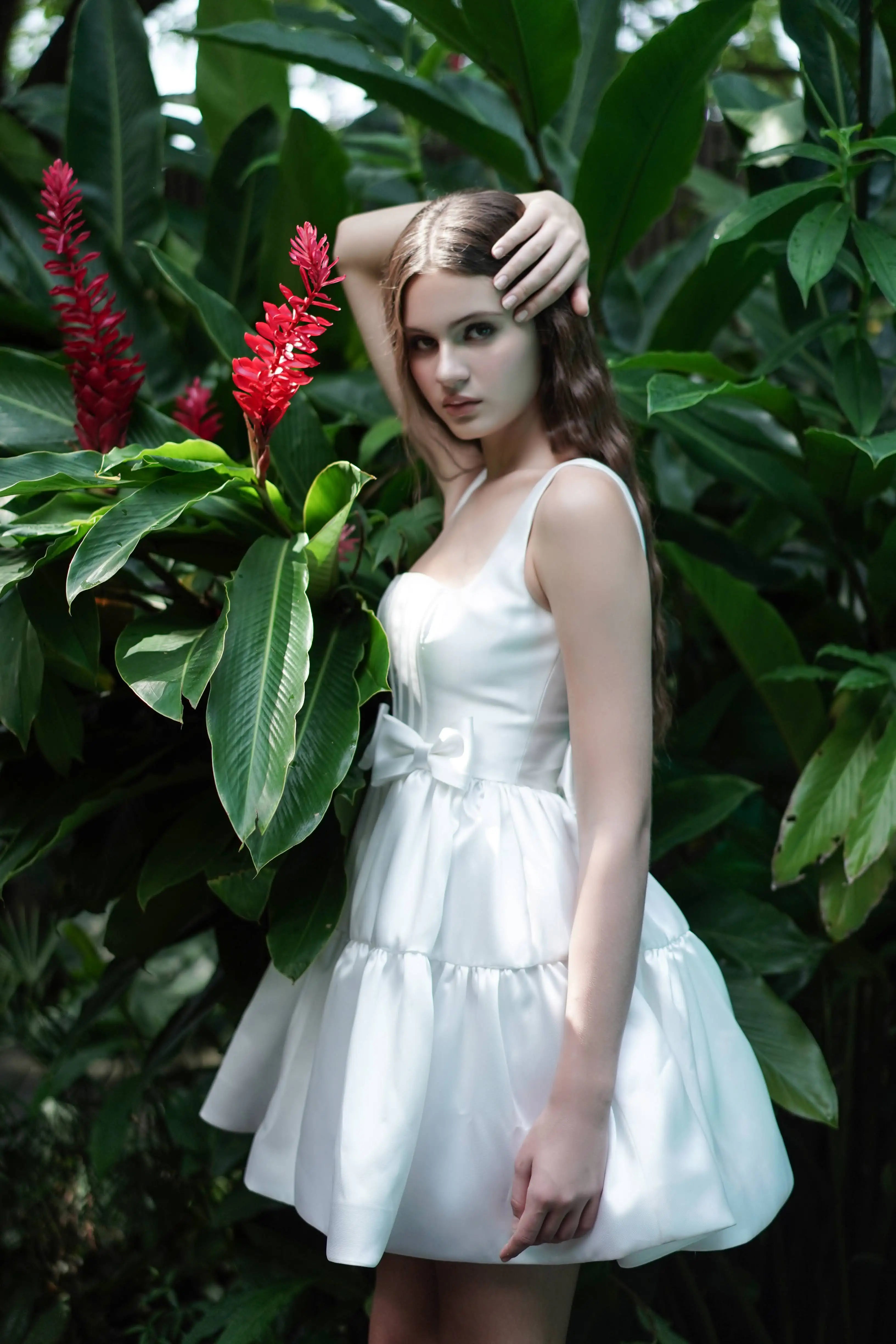 A young woman in the TH&TH Bloom Bridal Mini Dress in Ivory, featuring a corseted bodice and sleeveless satin twill design, stands among green foliage, gently holding a branch with red tropical flowers in natural light.