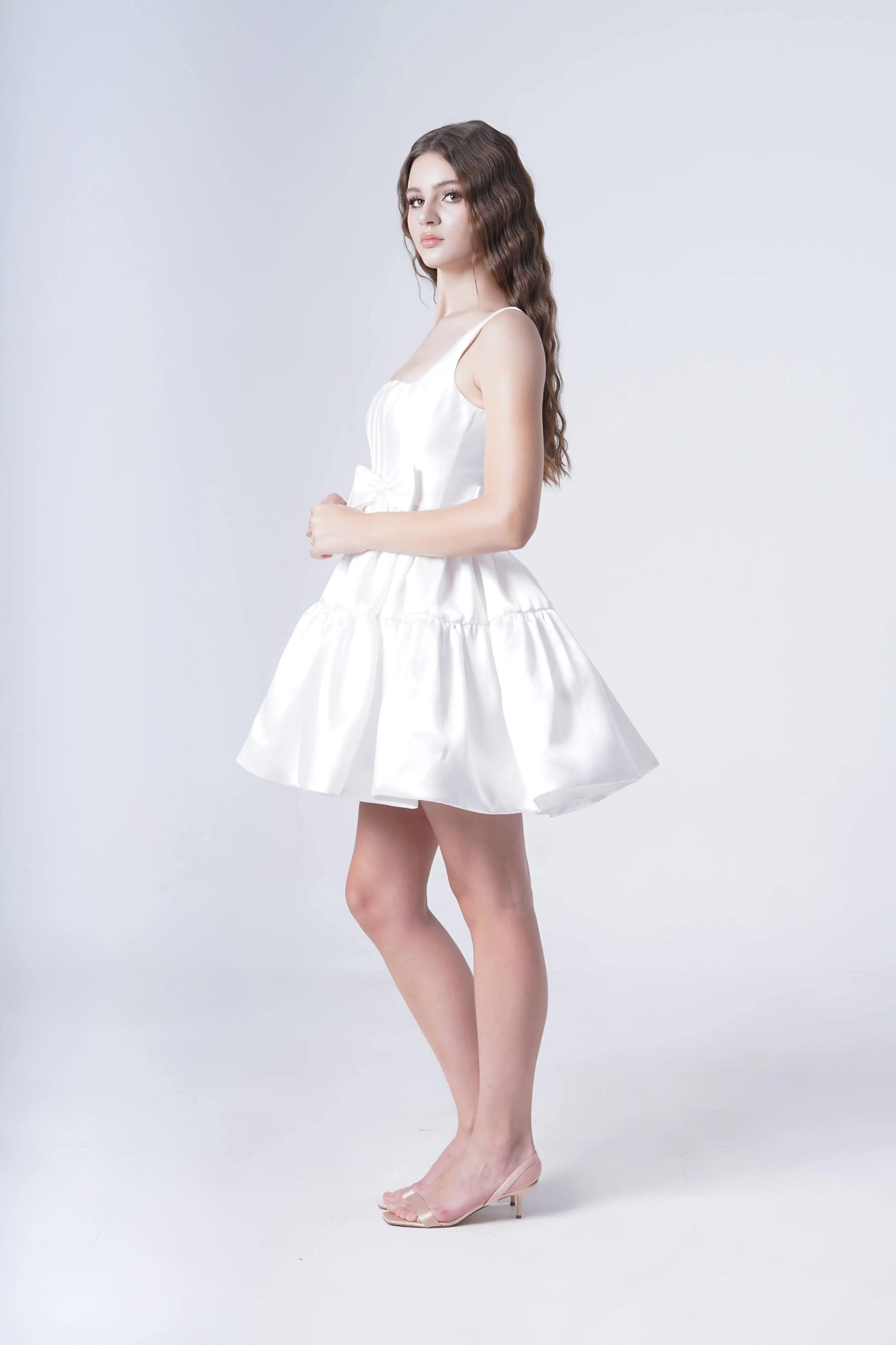 A young woman with long wavy hair models the TH&TH Bloom Bridal Mini Dress in Ivory, featuring a corseted bodice and sleeveless mini cut, paired with clear heels as she poses sideways against a plain white background.