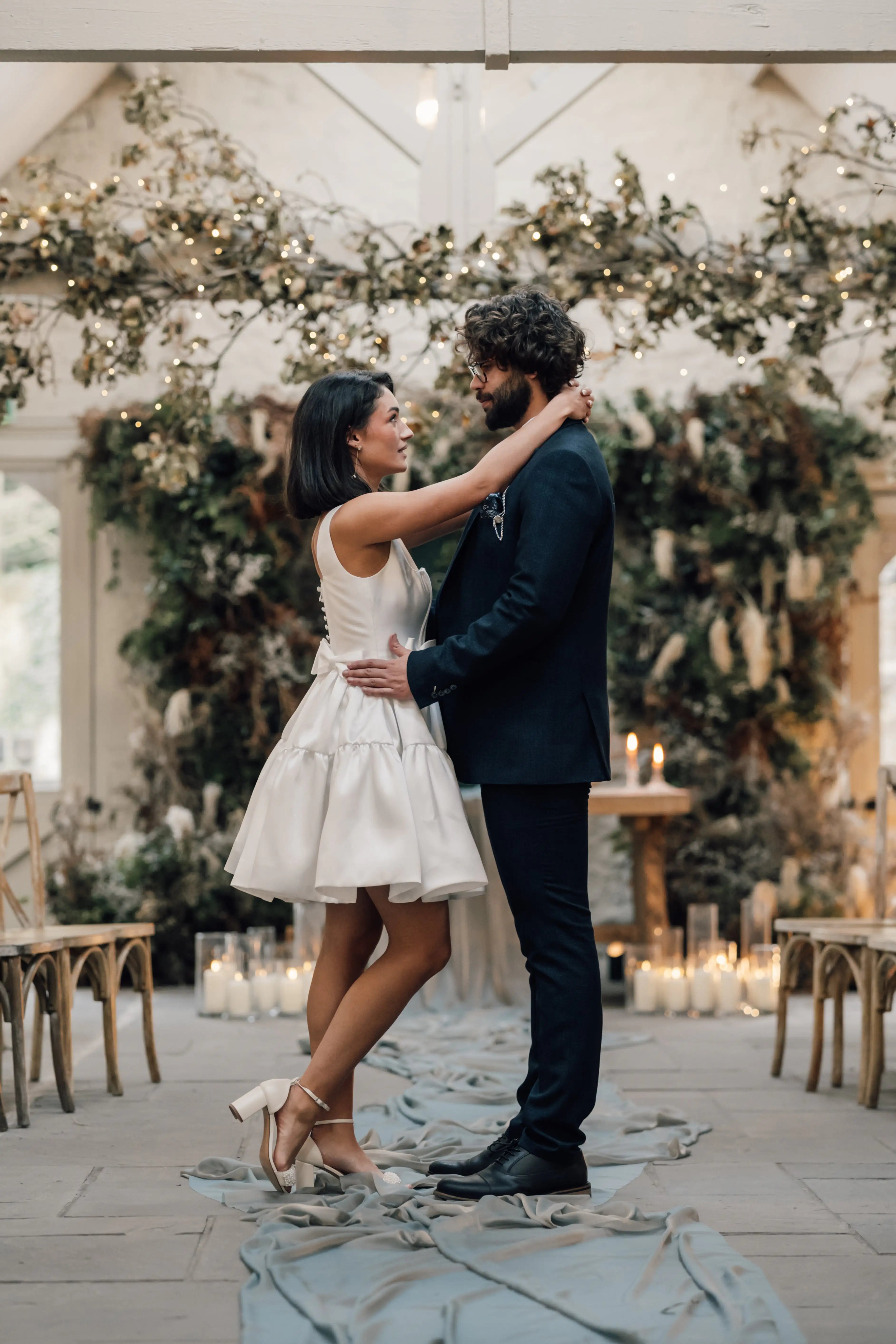 A couple stands close together, smiling at each other in a lushly decorated indoor venue with greenery, candles, and fairy lights. She wears the TH&TH Bloom Bridal Mini Dress in Ivory with a corseted bodice; he wears a dark suit.