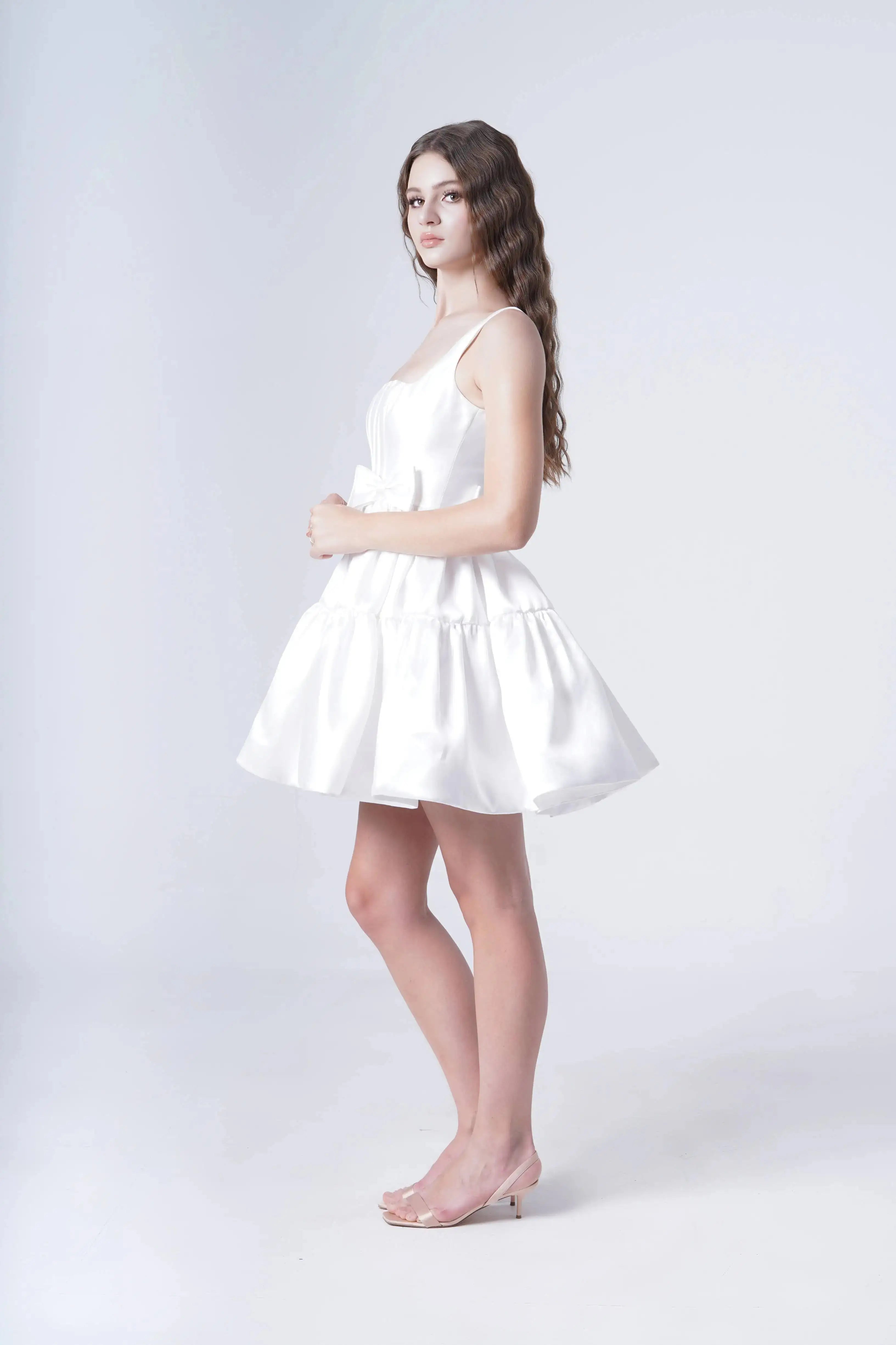 A young woman with long wavy hair models the TH&TH Bloom Bridal Mini Dress in Ivory, featuring a corseted bodice and sleeveless mini cut, paired with clear heels as she poses sideways against a plain white background.