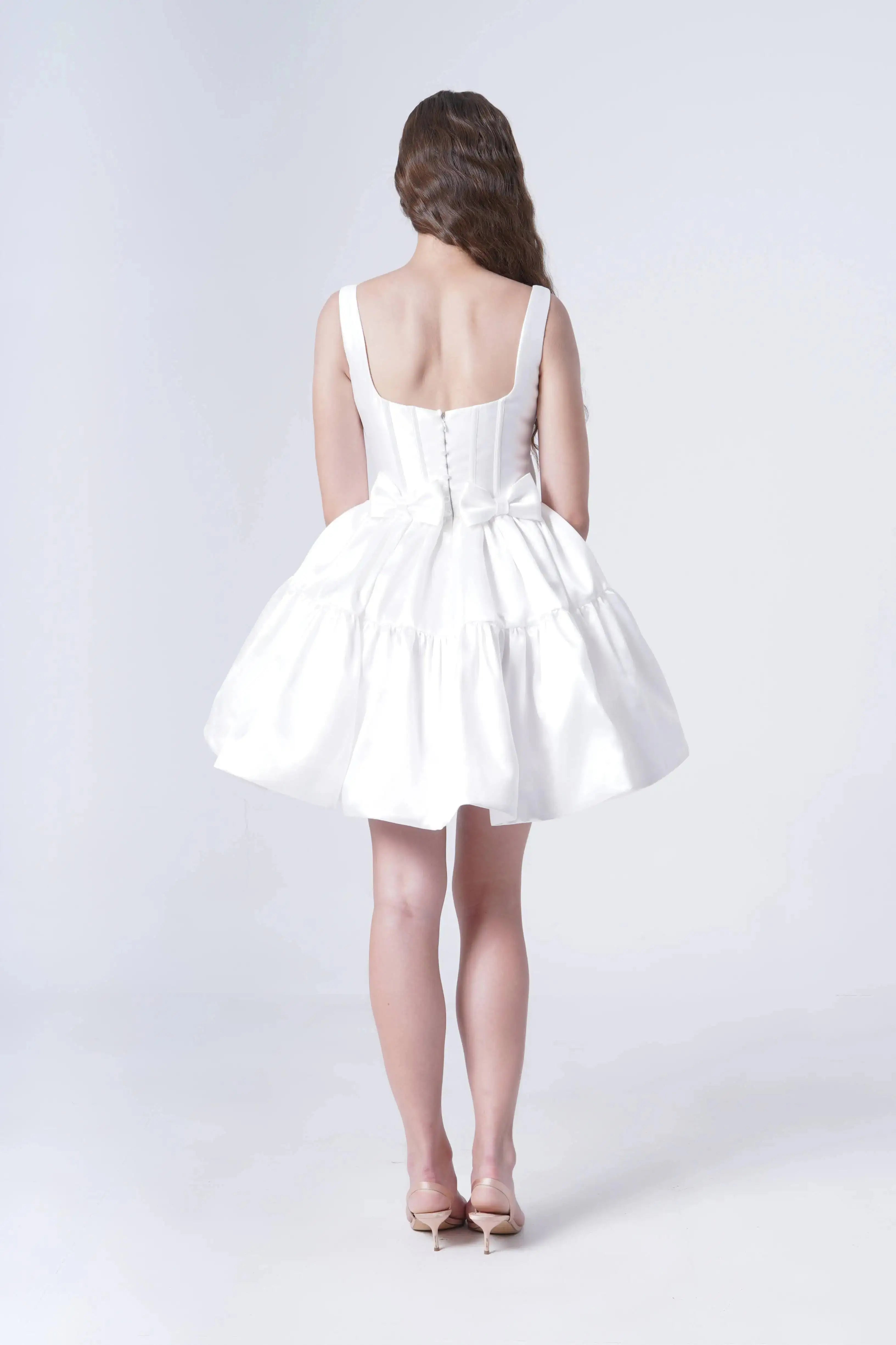 A woman with long brown hair stands with her back to the camera in the TH&TH Bloom Bridal Mini Dress in Ivory, featuring a flared skirt, corseted bodice, and waist bow, styled with nude heels against a plain light background.