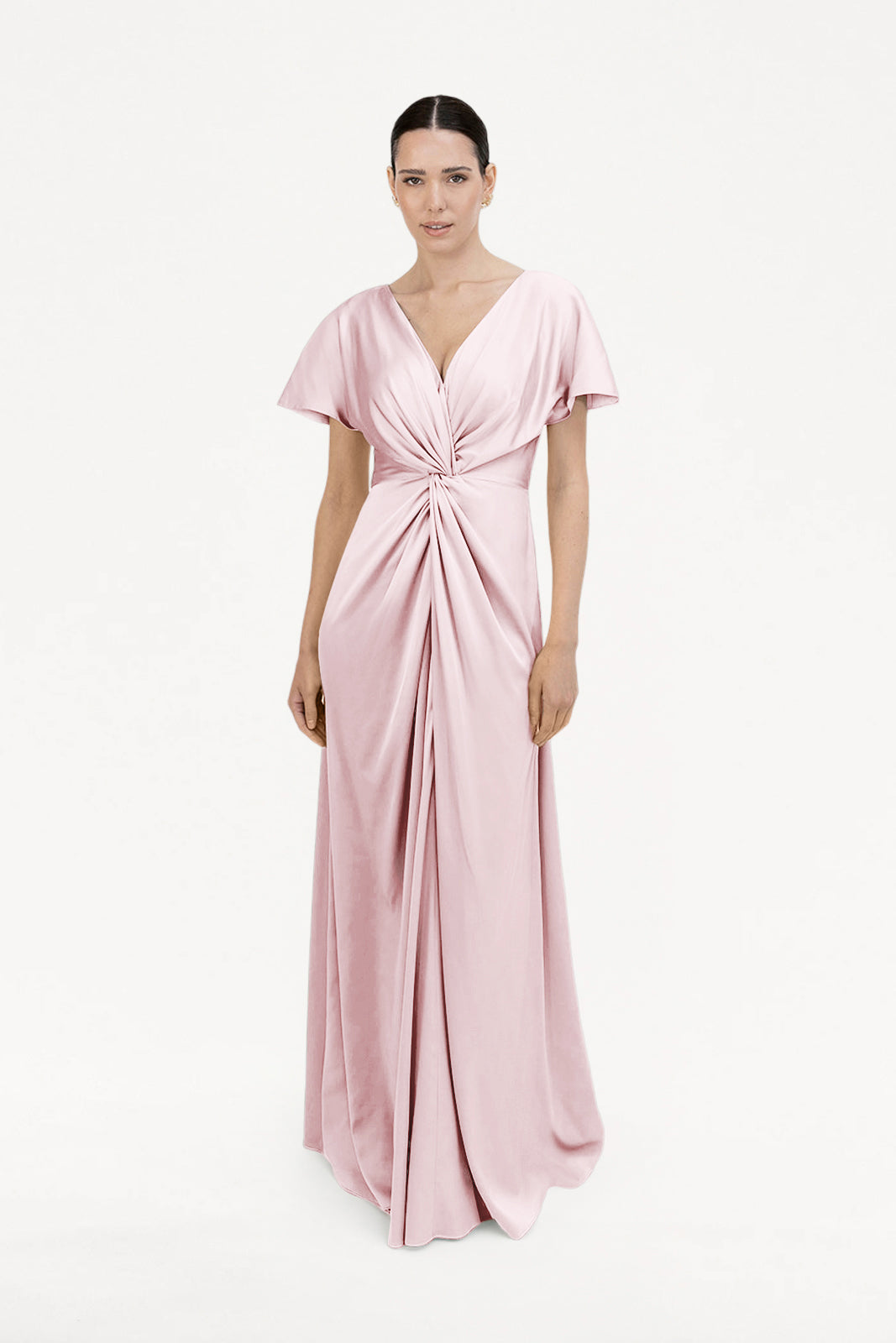 A woman models the Camilla Bridesmaid Dress in Blush Pink Satin by TH&TH, a floor-length gown with short sleeves and a twisted front detail—an elegant, modern bridesmaid choice—against a plain white background.