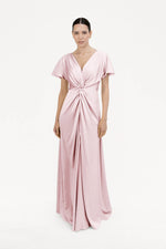 A woman models the Camilla Bridesmaid Dress in Blush Pink Satin by TH&TH, a floor-length gown with short sleeves and a twisted front detail—an elegant, modern bridesmaid choice—against a plain white background.